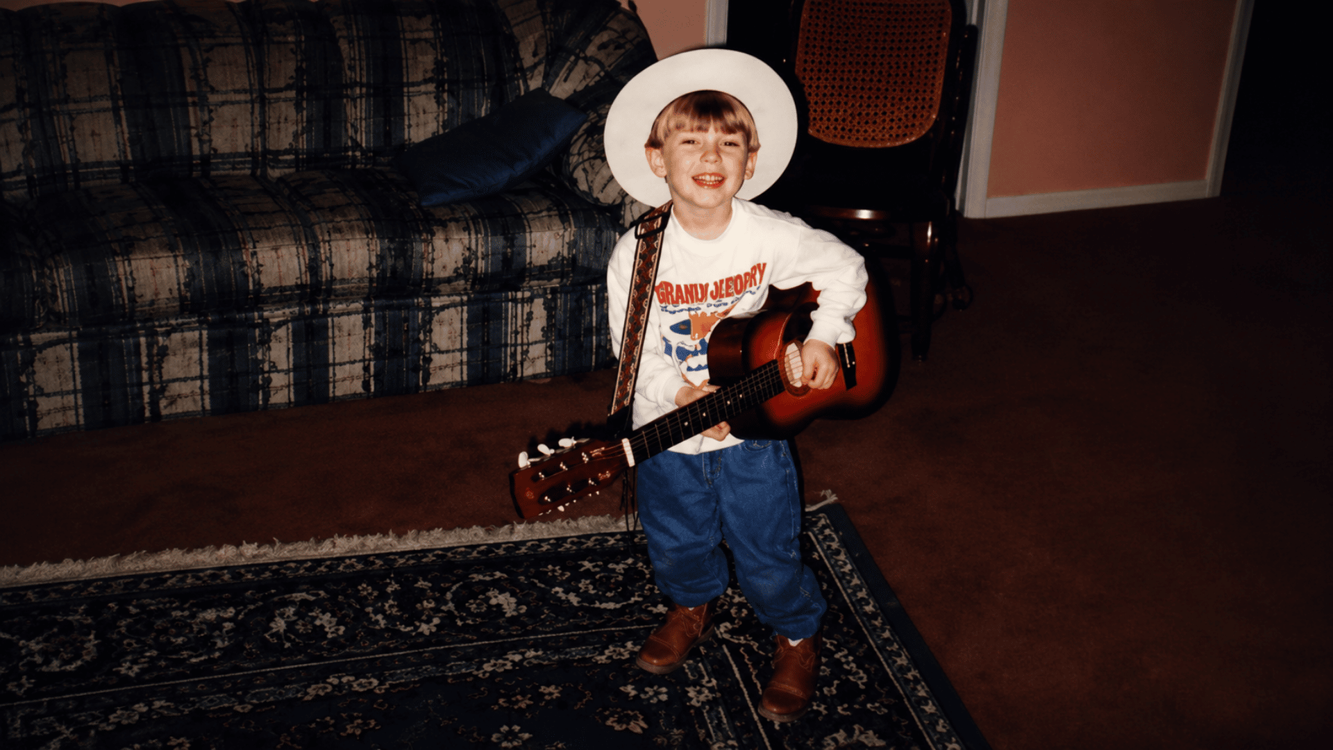 Childhood photo of Morgan Wallen smiling in a cowboy hat while holding an acoustic guitar inside a living room.