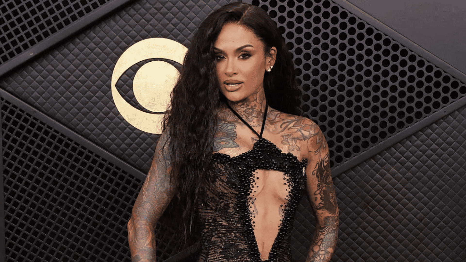 kehlani posing in a black dress against a dark patterned wall with a gold emblem