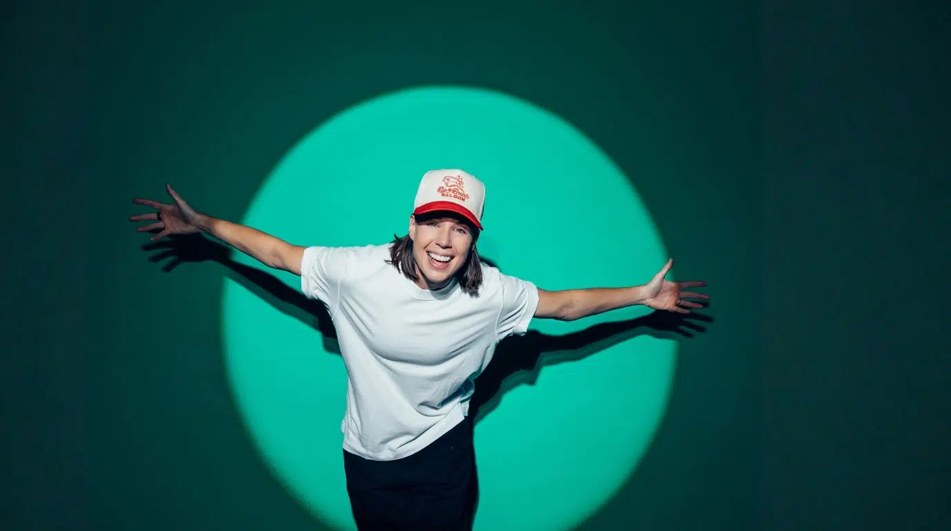 person wearing a white shirt and cap posing with arms extended under a spotlight backdrop