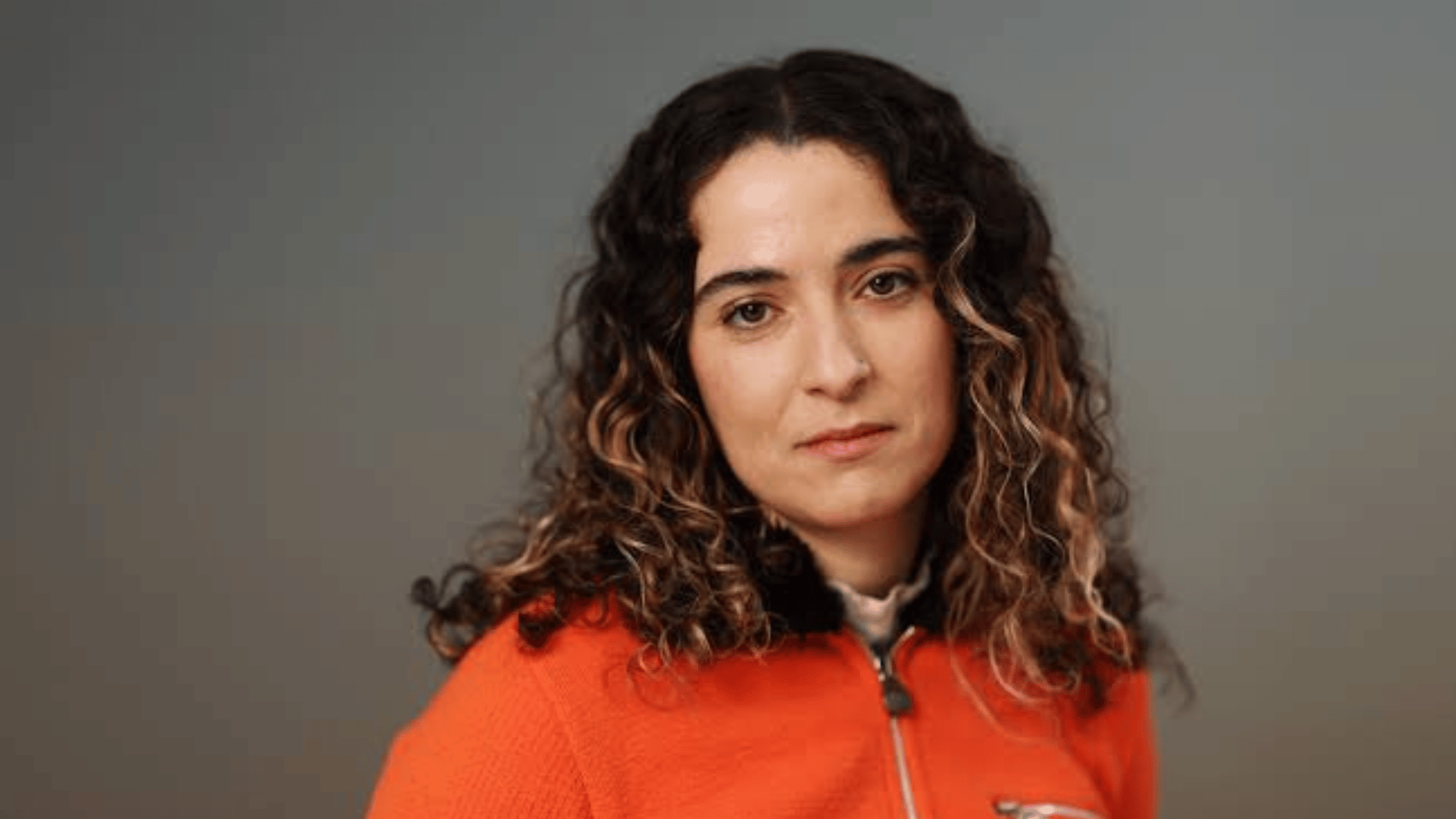 rachel kaly with curly dark hair wearing a bright orange zip up jacket against a neutral gray background