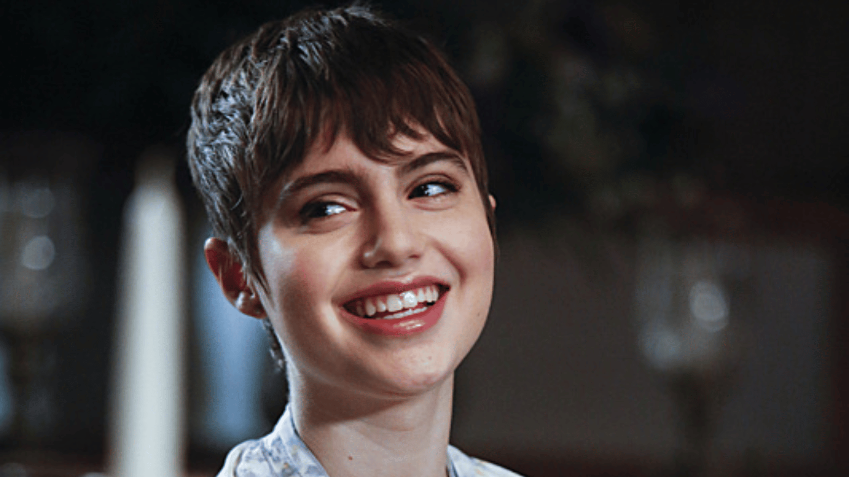sami gayle with short brown hair smiles while looking to the side, shown in a close-up portrait with a dark blurred background