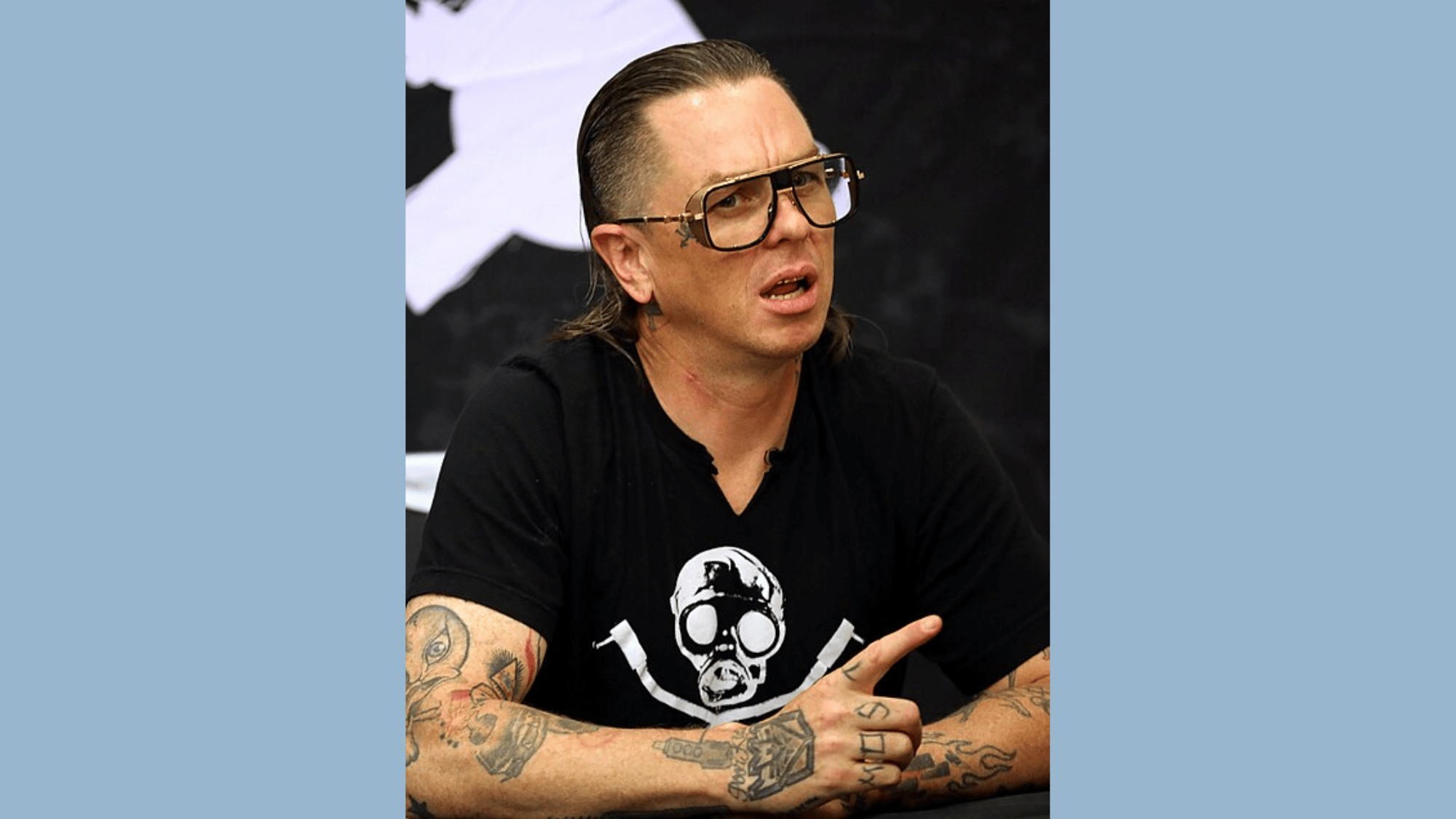 sid wilson seated at a table wearing large glasses and a black graphic shirt with tattoos visible across both forearms