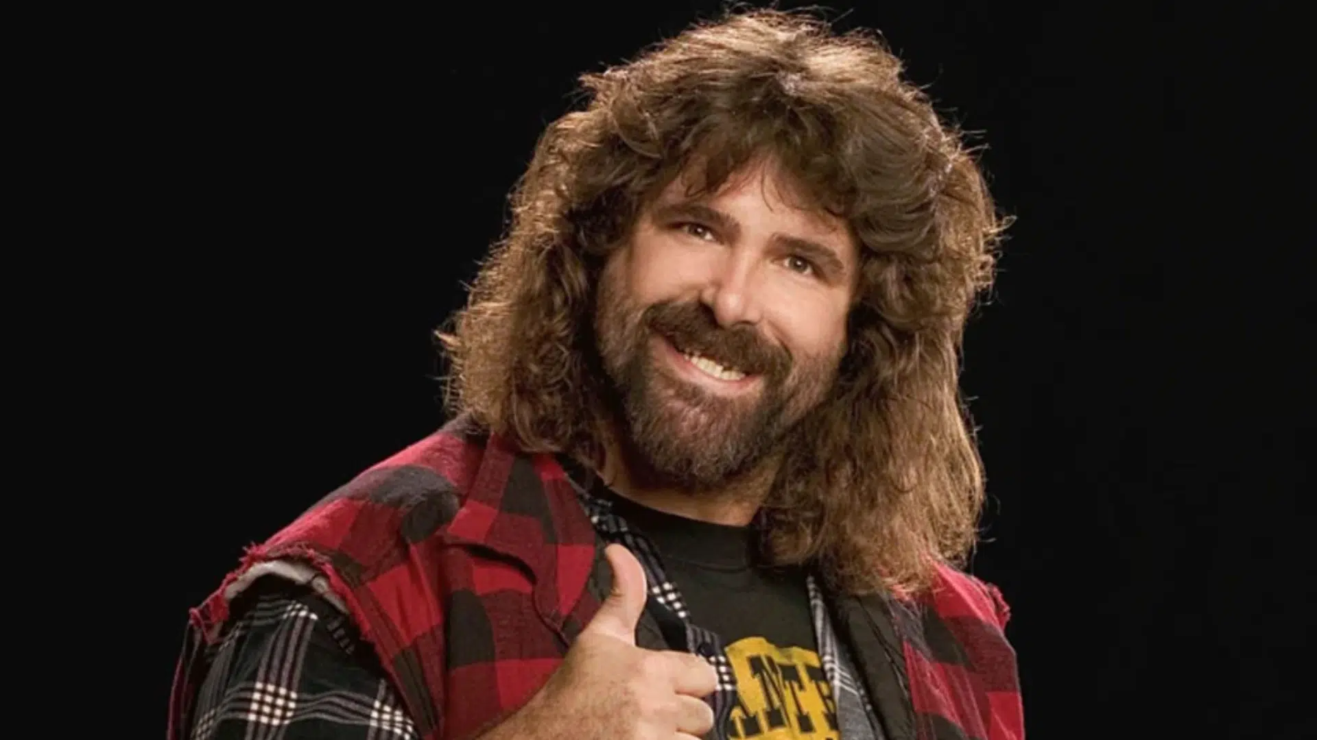 smiling bearded wrestler with long brown hair wearing a red plaid vest and giving a thumbs up against black background