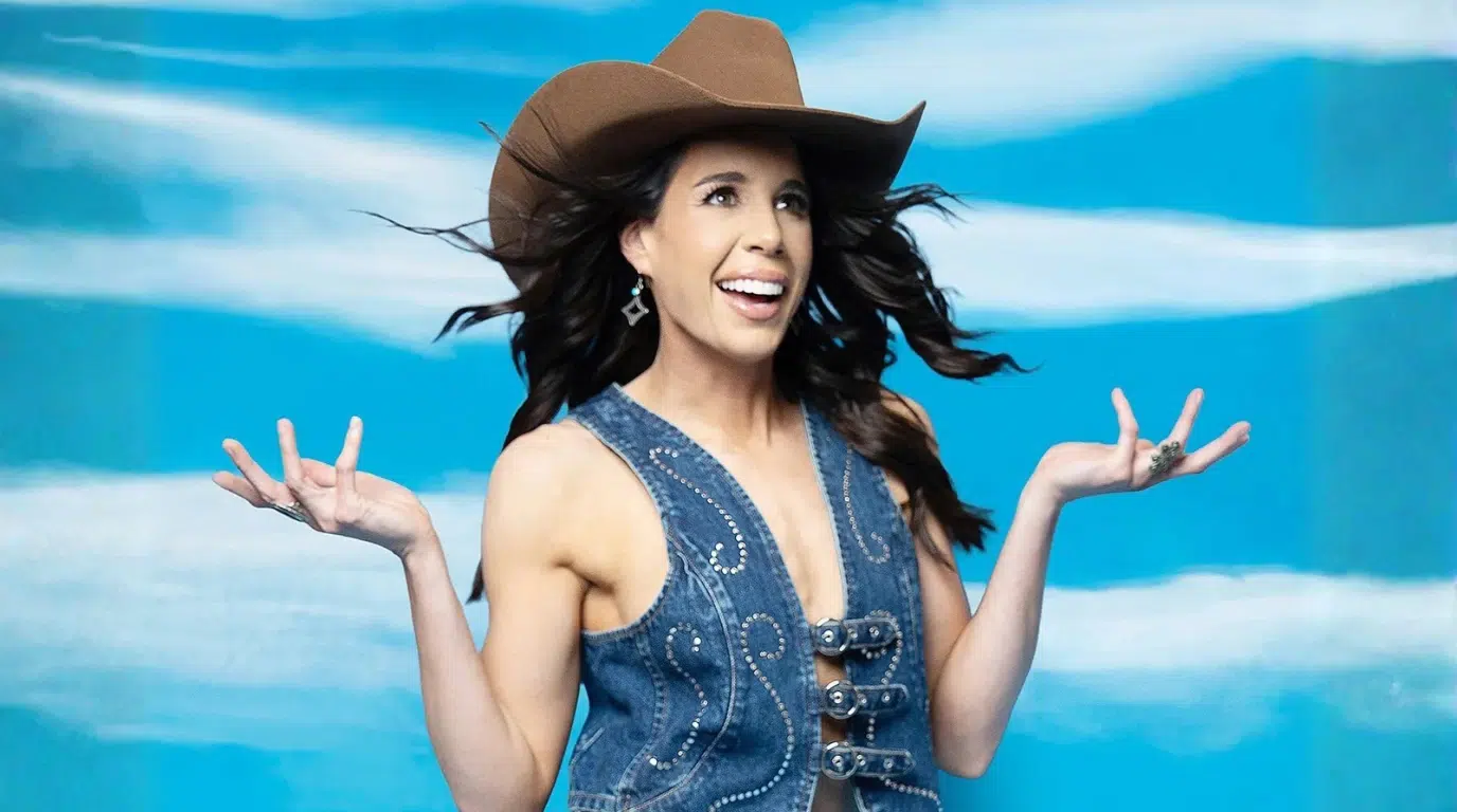 smiling woman wearing a brown cowboy hat and denim western vest posing with raised hands against a blue background