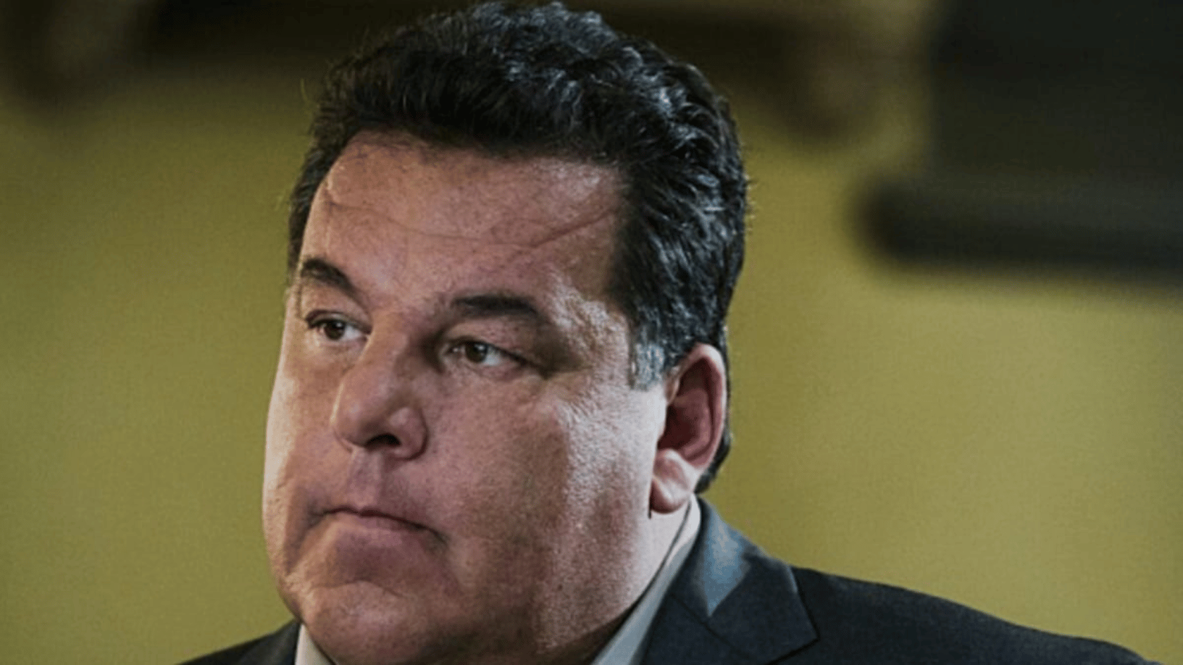 steve schirripa with dark hair wearing a suit looks to the side with a serious expression in an indoor setting with a blurred background