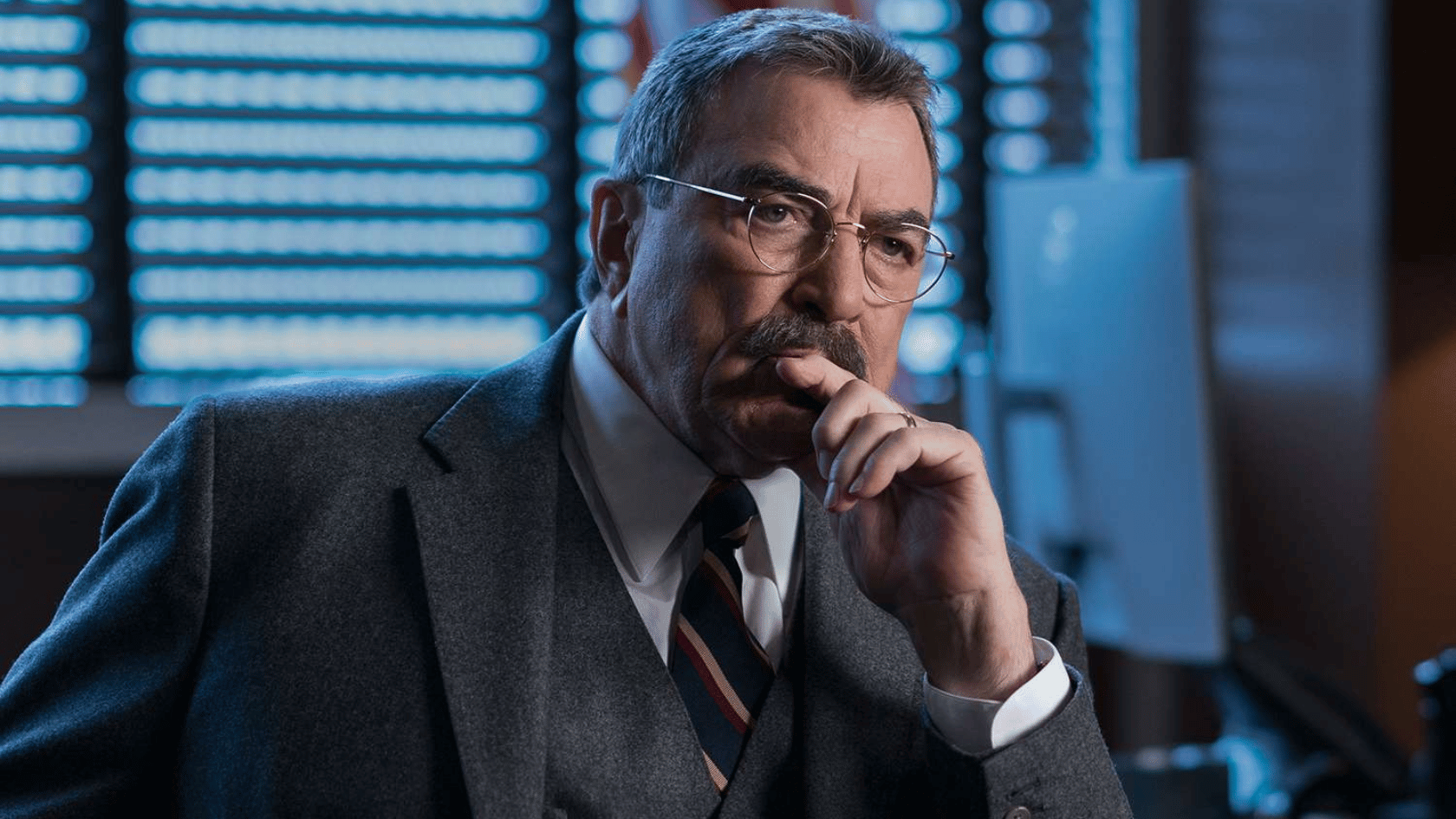 tom selleck in a gray suit and glasses sits in an office, resting his hand near his face while looking thoughtful under cool blue lighting