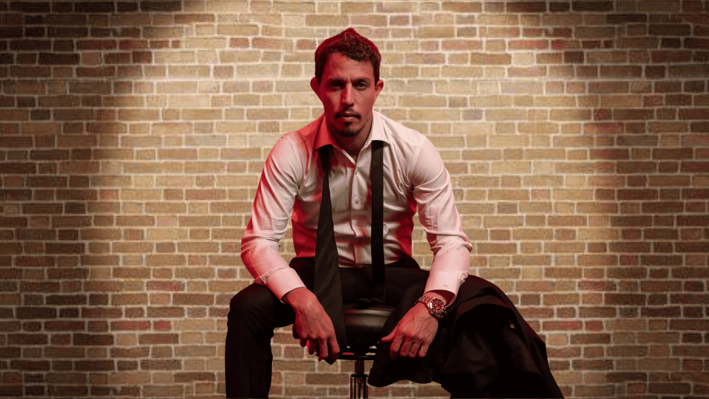 tony hinchcliffe seated in a white shirt and black tie against a brick wall