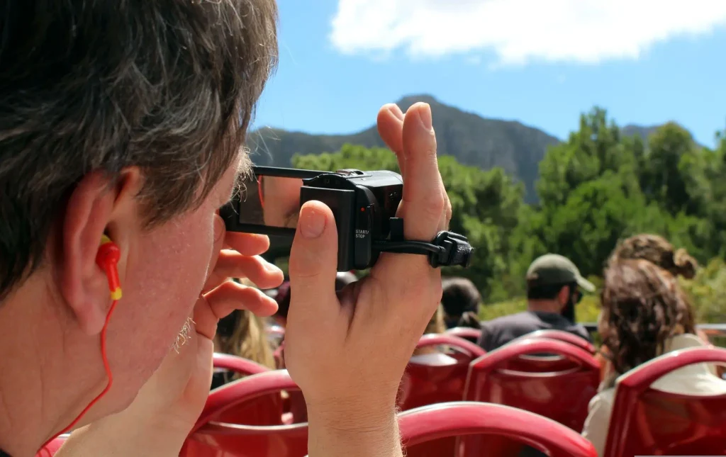 Person filming with a camcorder at an outdoor event with red chairs and trees