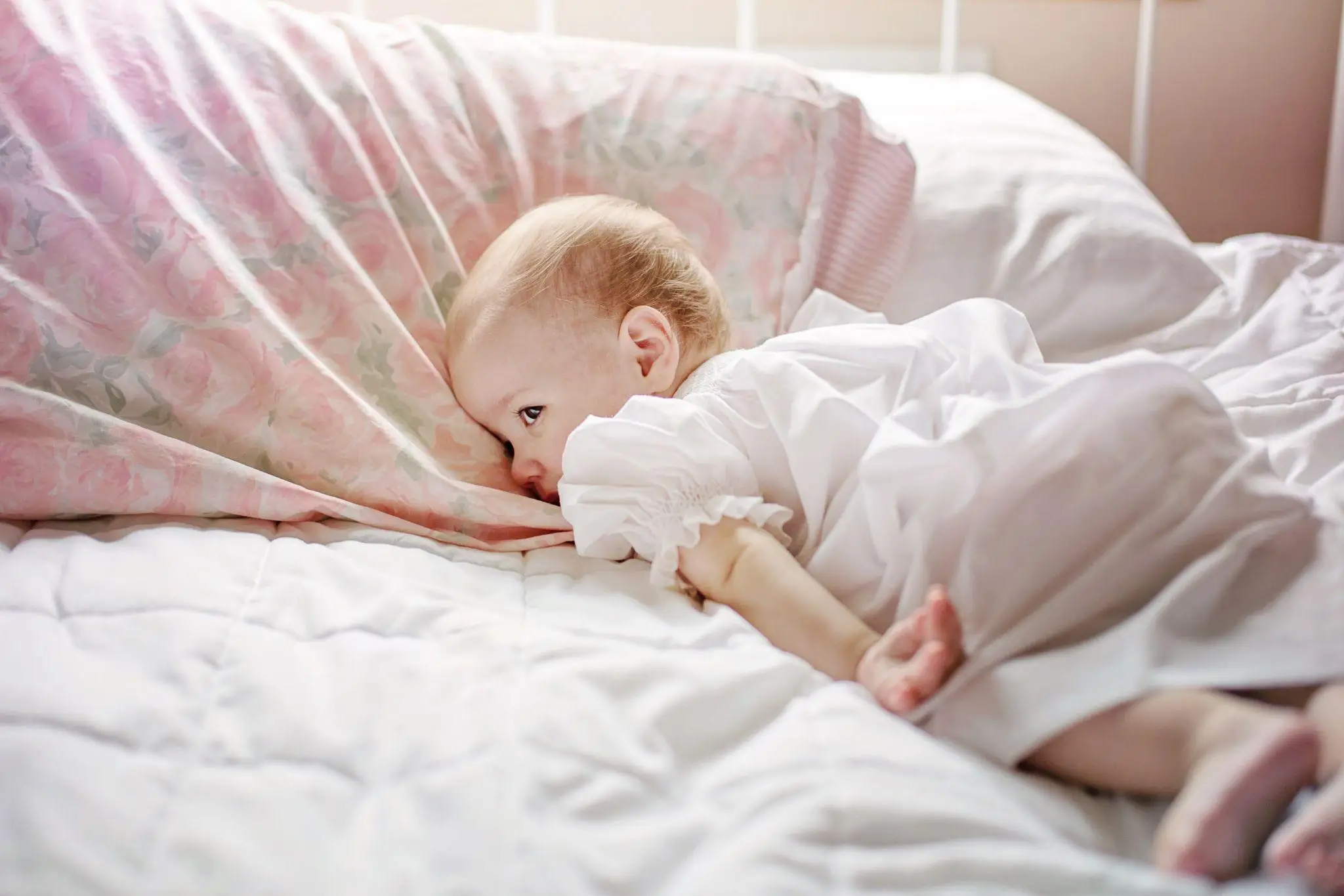 Baby in white dress lying on bed with floral pillow in soft daylight