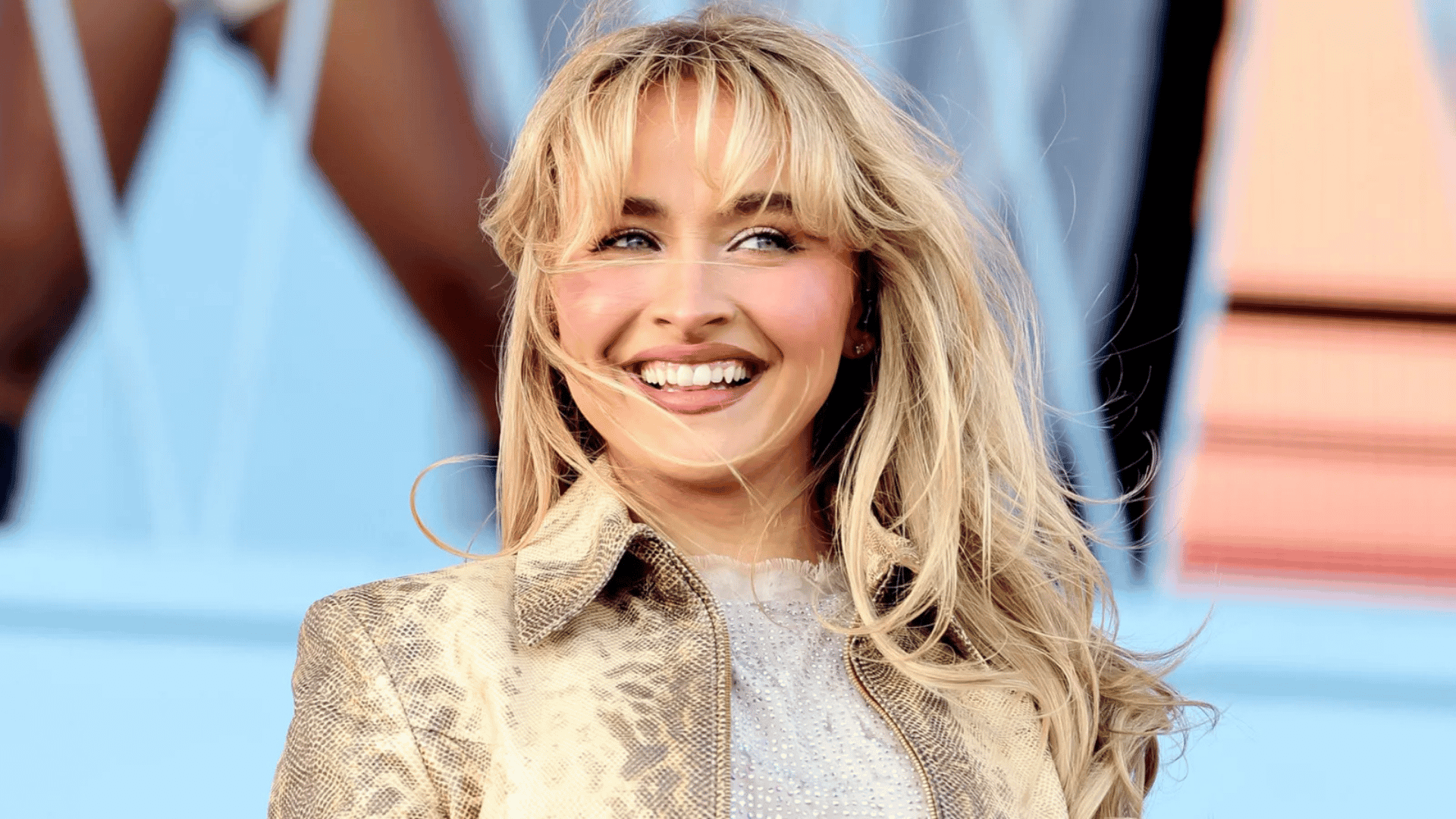 sabrina carpenter smiling outdoors in a patterned jacket against a blue backdrop
