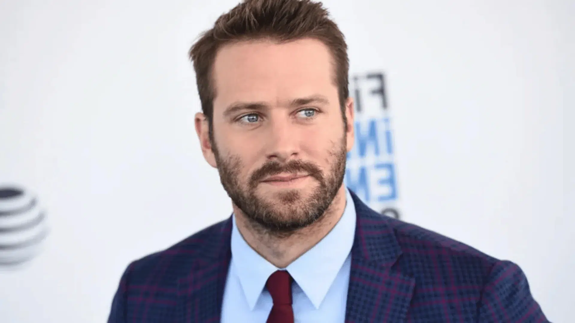 armie hammer wearing a plaid blazer and red tie looking to the side at a public event