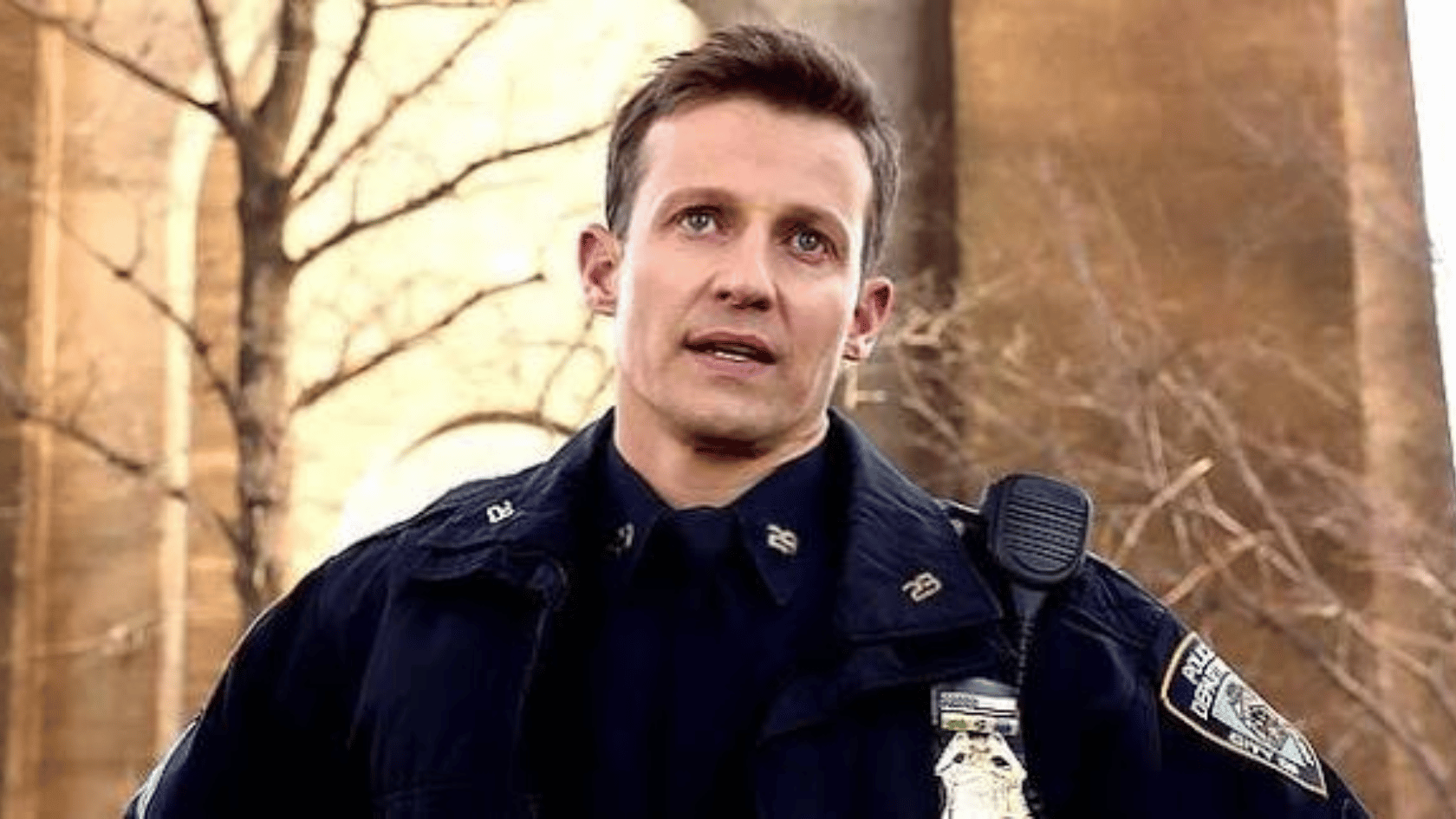 will estes wearing a dark police uniform stands outdoors near a building, looking ahead with a serious expression