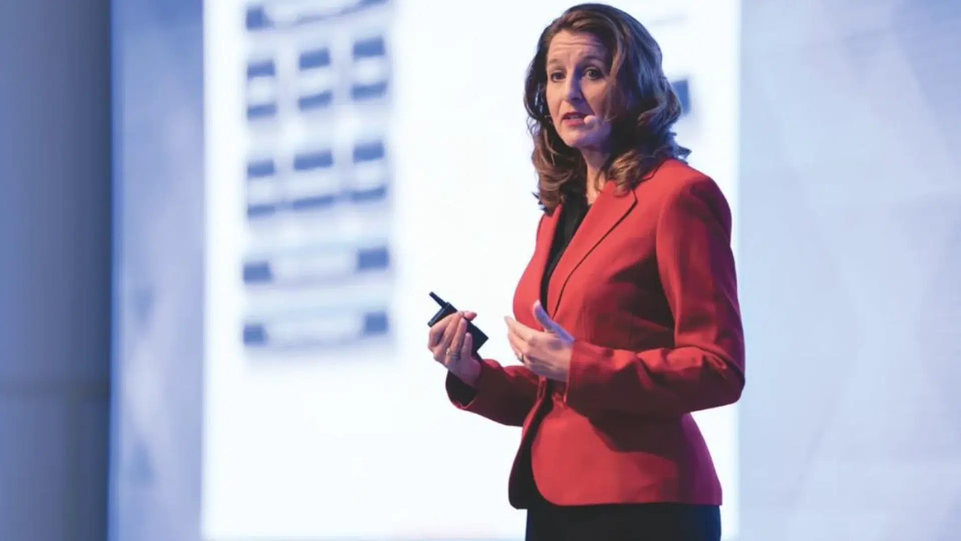 woman in red blazer speaking on stage with headset microphone holding a clicker against a blurred presentation screen