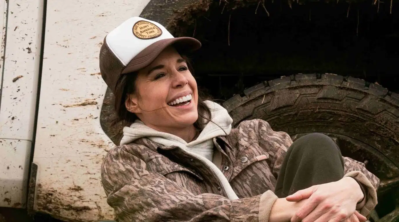 woman wearing a cap and camouflage jacket smiling while sitting near a vehicle outdoors