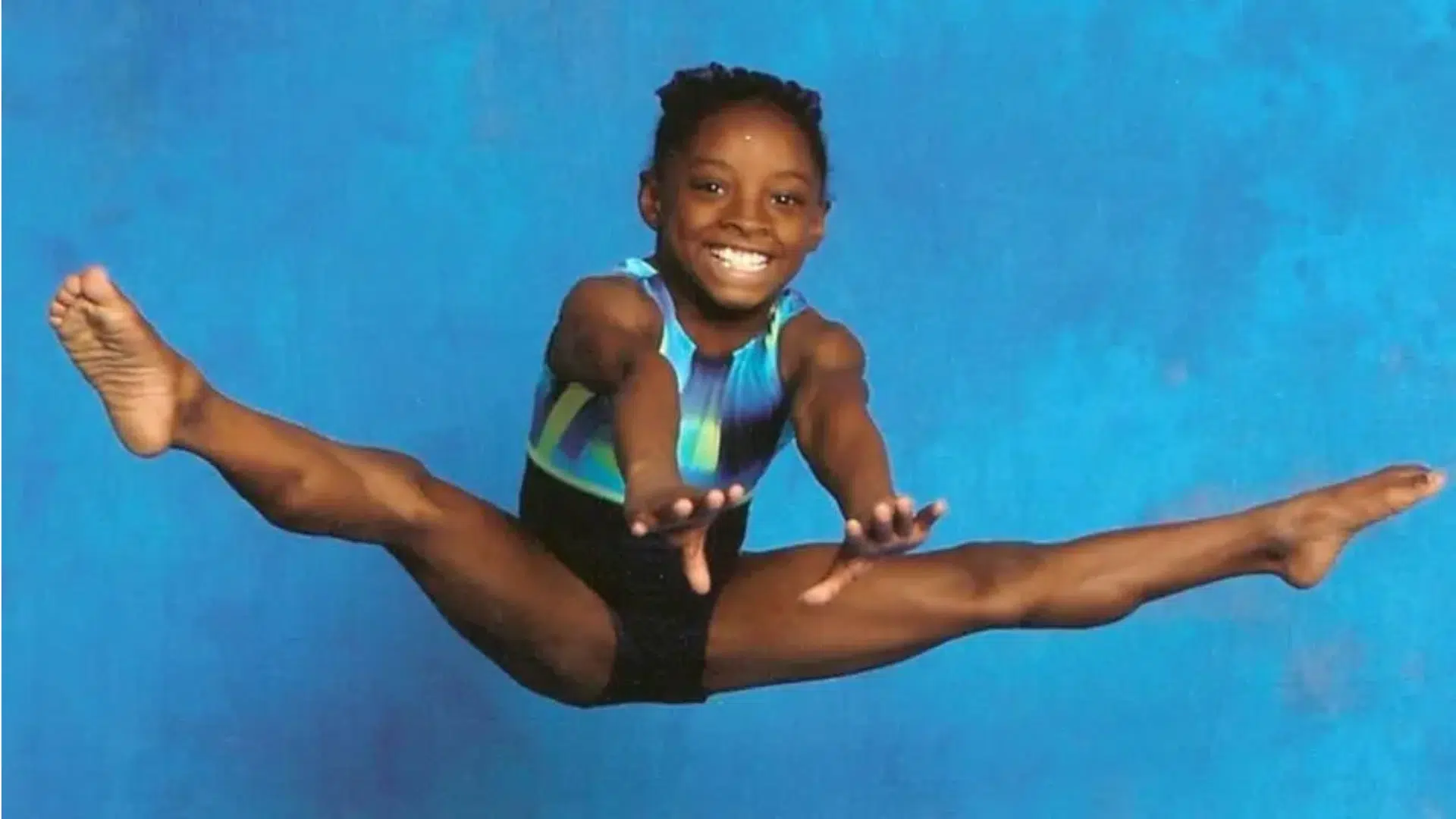young gymnast performing a split leap in midair against a blue background with a bright smile