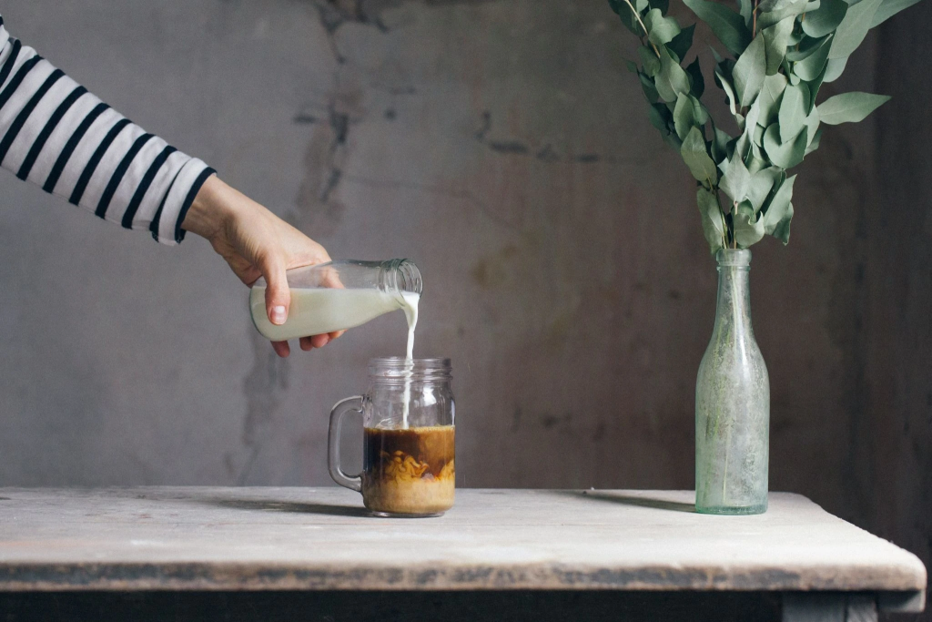 Pouring milk into a mason jar of coffee on rustic table with green foliage in bottle