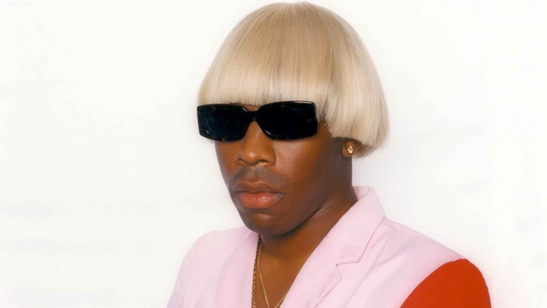 Tyler, The Creator wearing a blonde bowl-cut hairstyle, black sunglasses, and a pink suit jacket in a portrait.