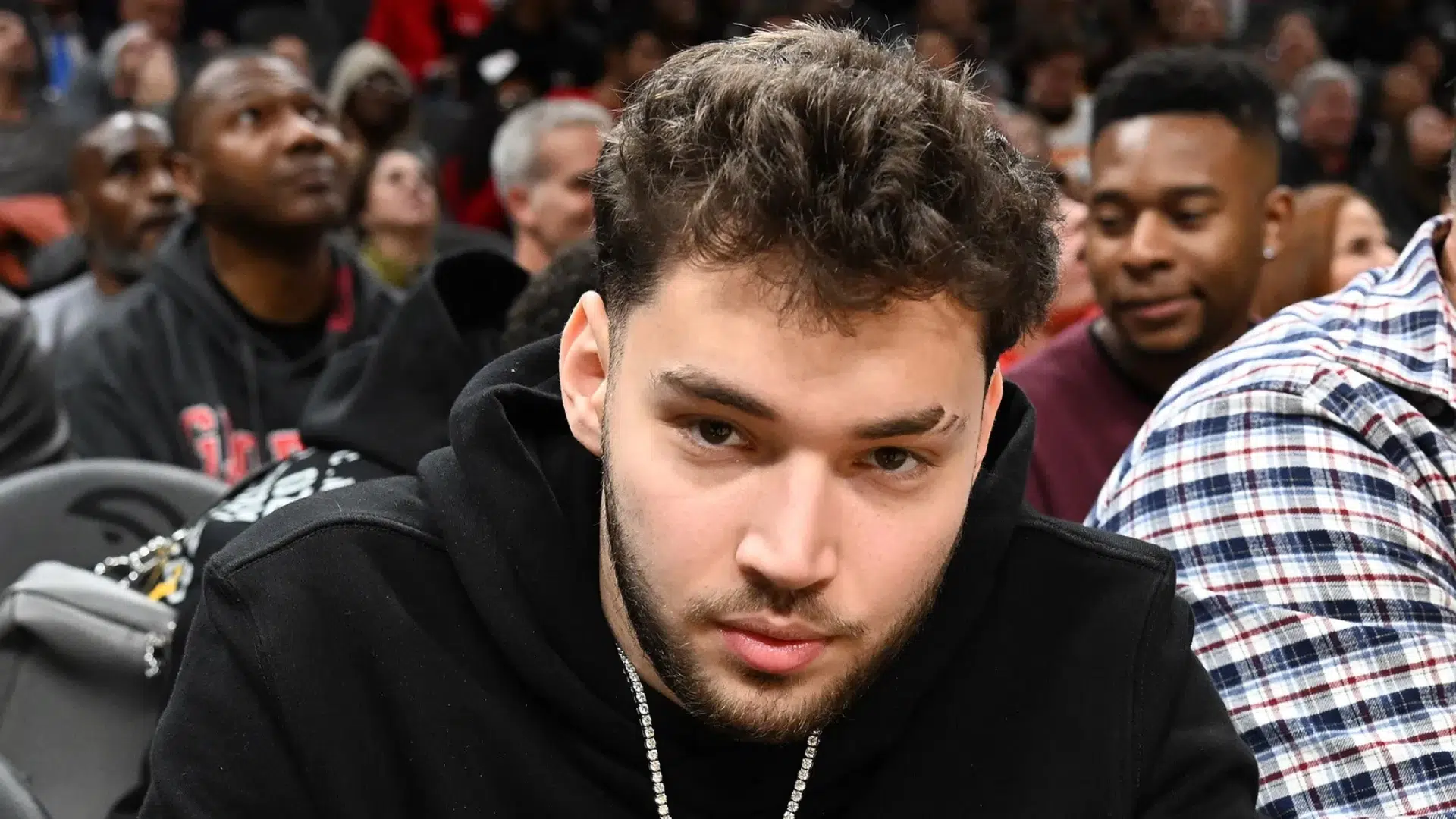 young male streamer in a black hoodie seated courtside at a crowded basketball game