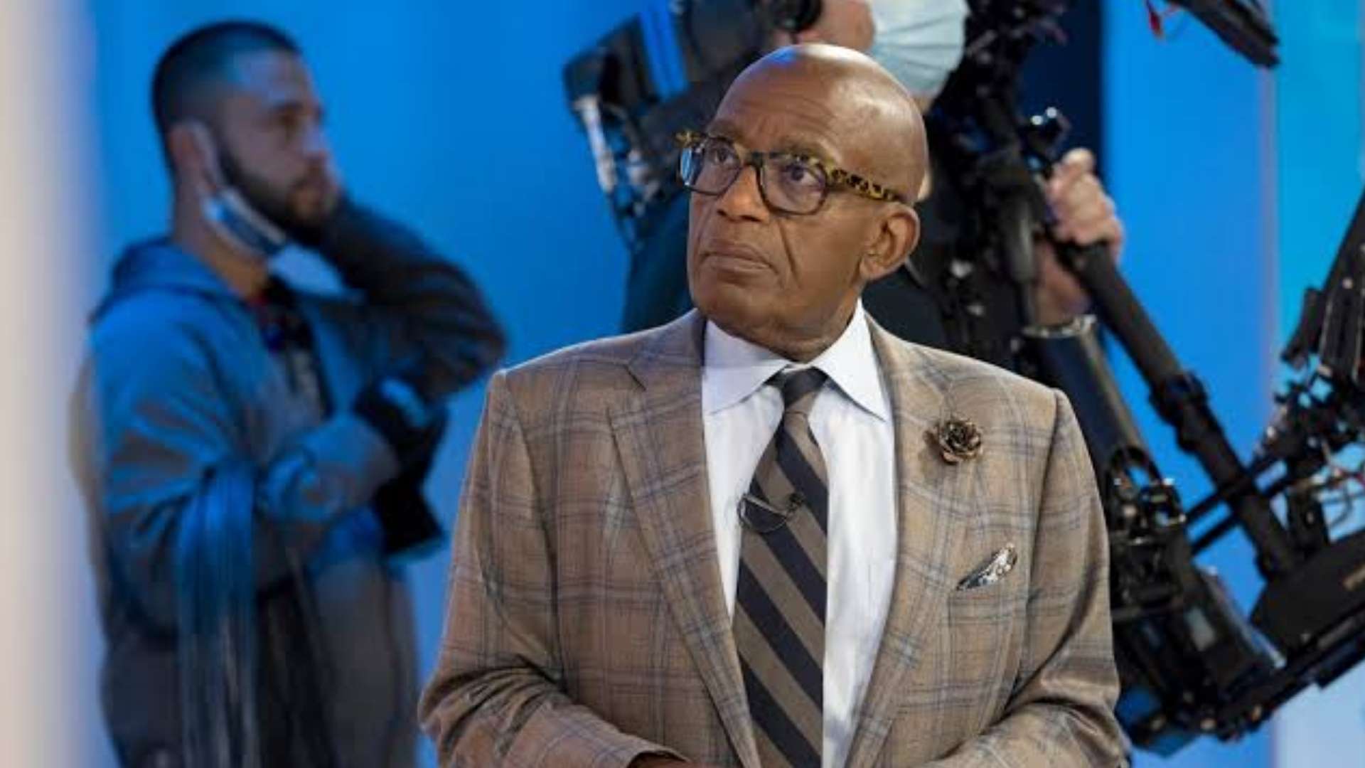 Al Roker looking upward while standing in a television studio in a plaid blazer and tie.