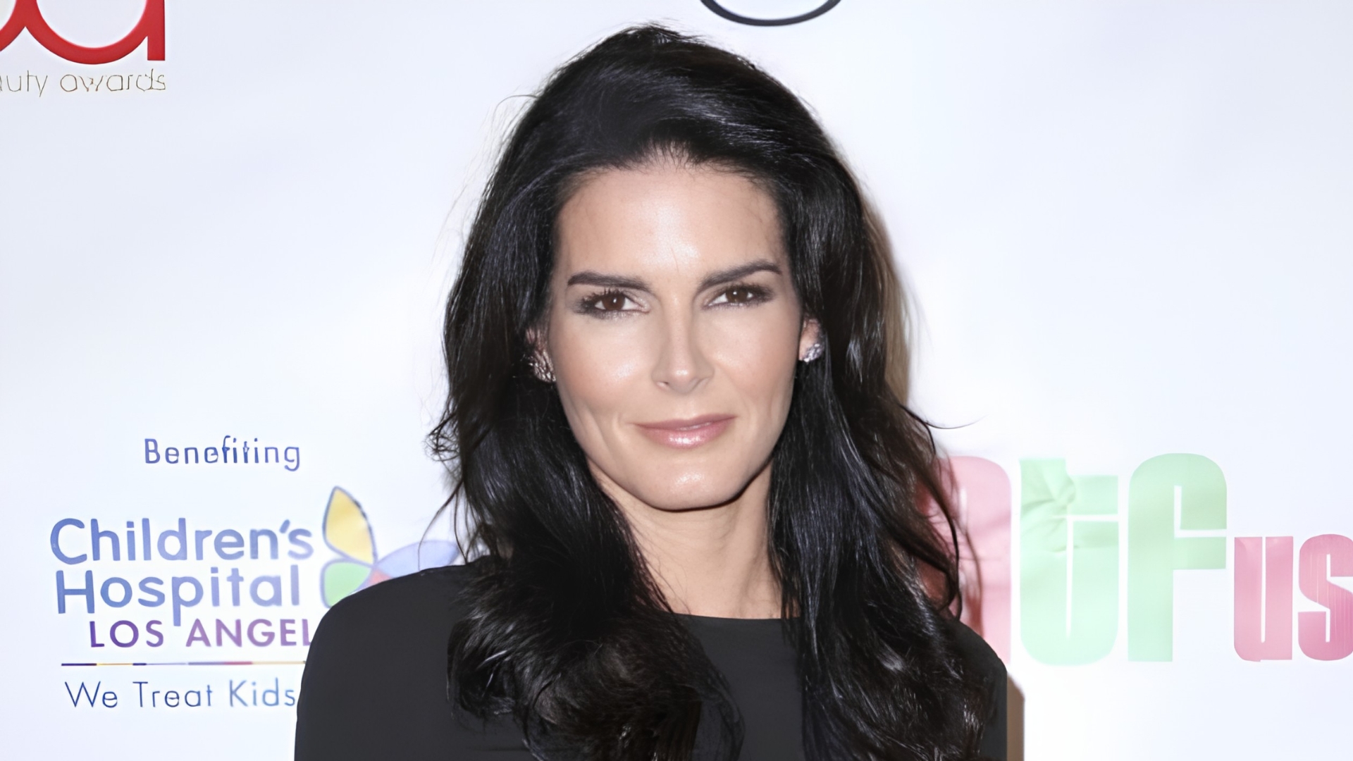 Angie Harmon with dark, wavy hair posing in a black top in front of a Children's Hospital Los Angeles backdrop.