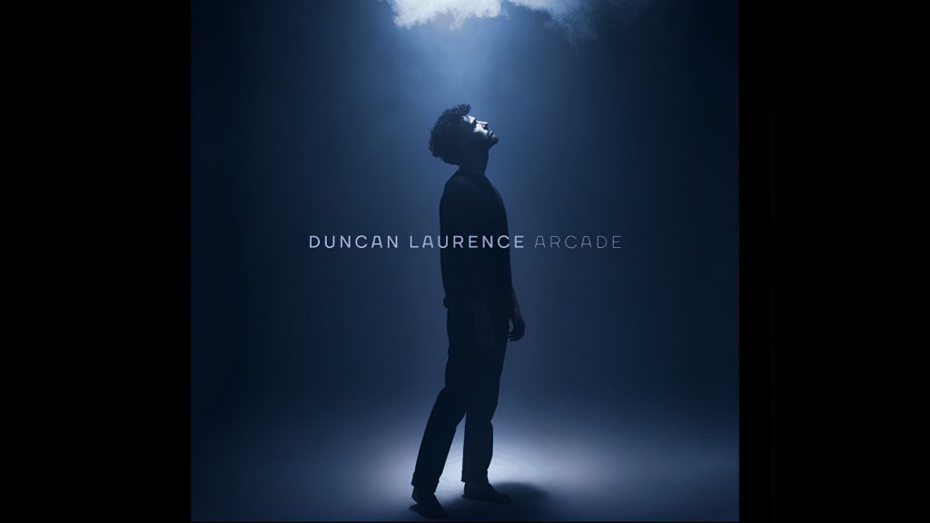 Duncan Laurence standing under a spotlight in a dark blue setting, representing the emotional atmosphere of his song “Arcade”