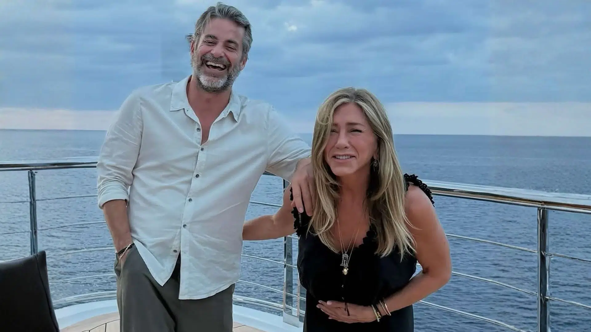 Jennifer Aniston and Jim Curtis smiling together on a boat deck with ocean background and casual vacation attire