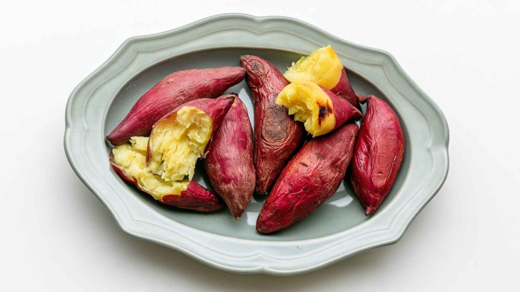 Roasted sweet potatoes with red skin served on a gray ceramic plate with a few pieces cut open showing the yellow flesh.