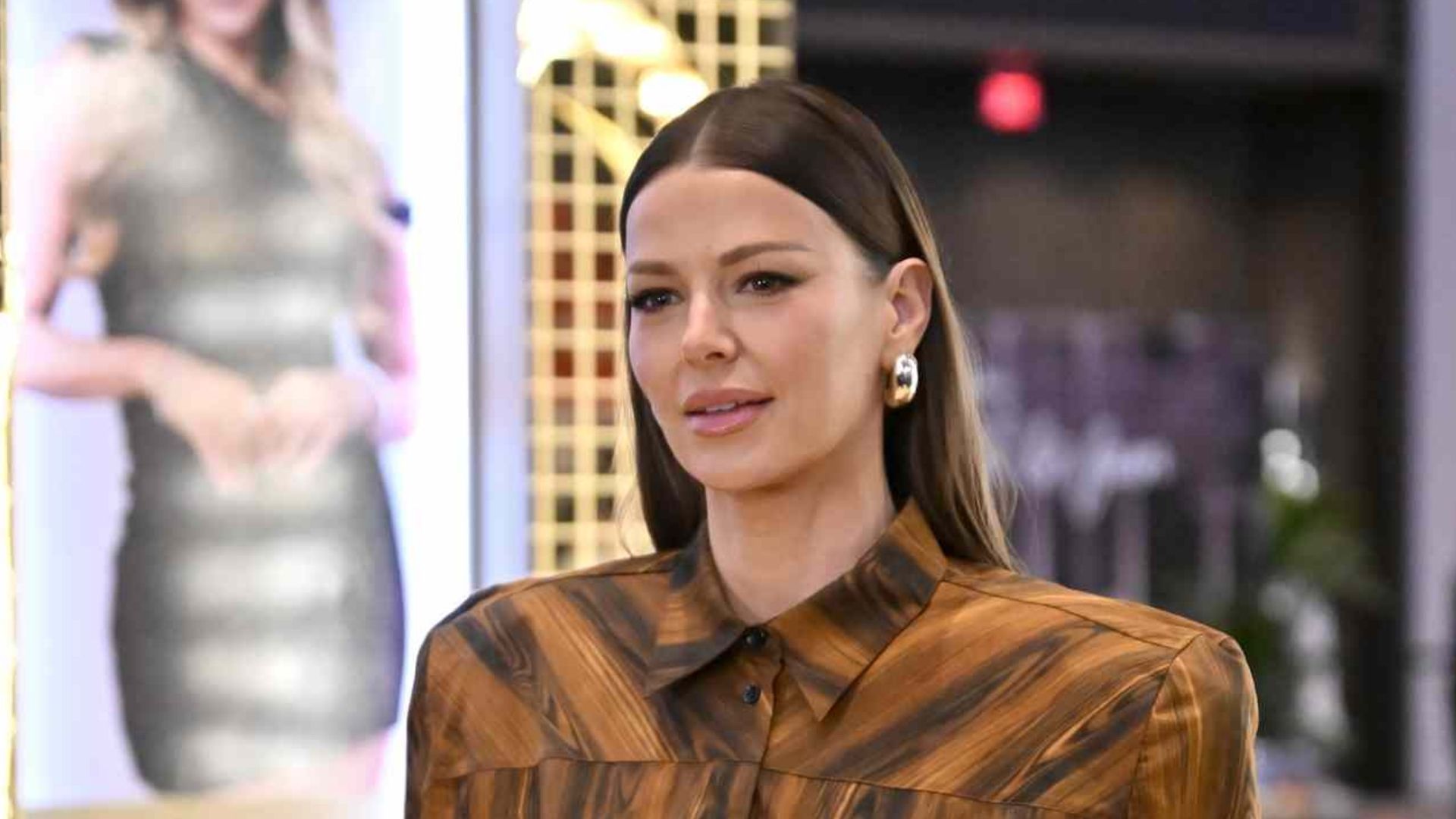 A woman walking forward wearing a matching brown wood-grain patterned outfit consisting of a structured, split-sleeve button-up shirt and wide-leg trousers, with a gold display booth in the background.