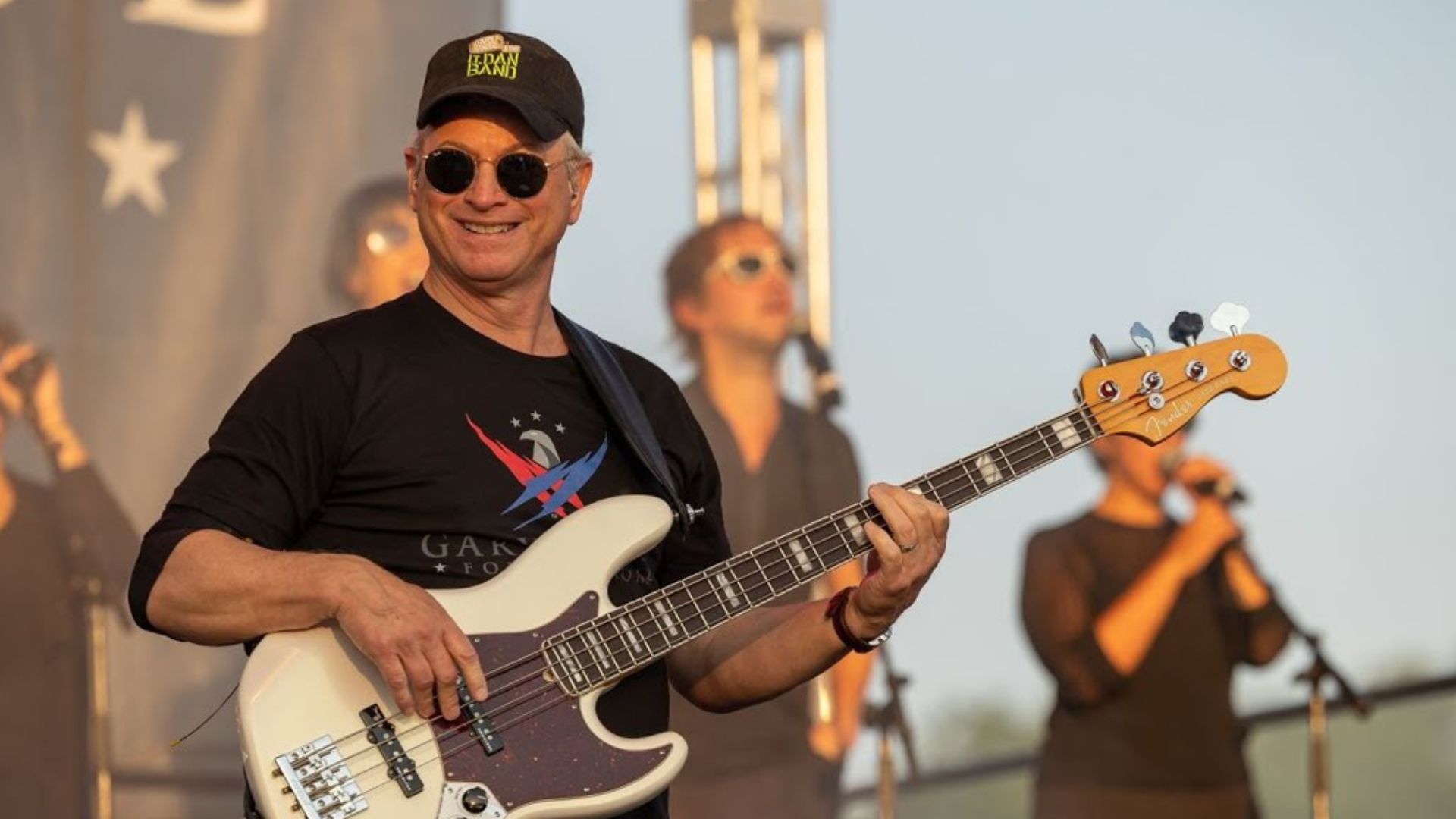 Gary Sinise playing bass guitar during a live stage performance with musicians in the background.