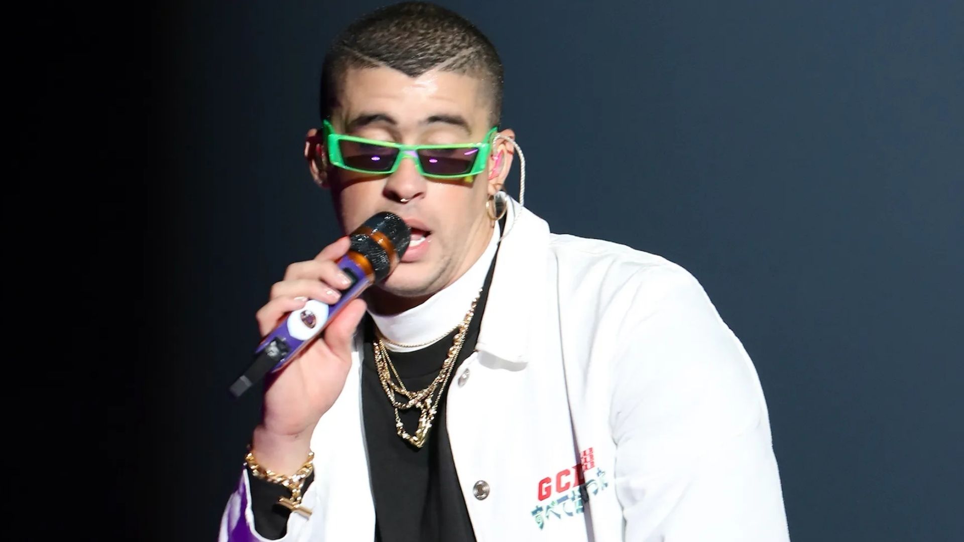 Bad Bunny performs onstage in green sunglasses and a white jacket while singing into a handheld microphone.