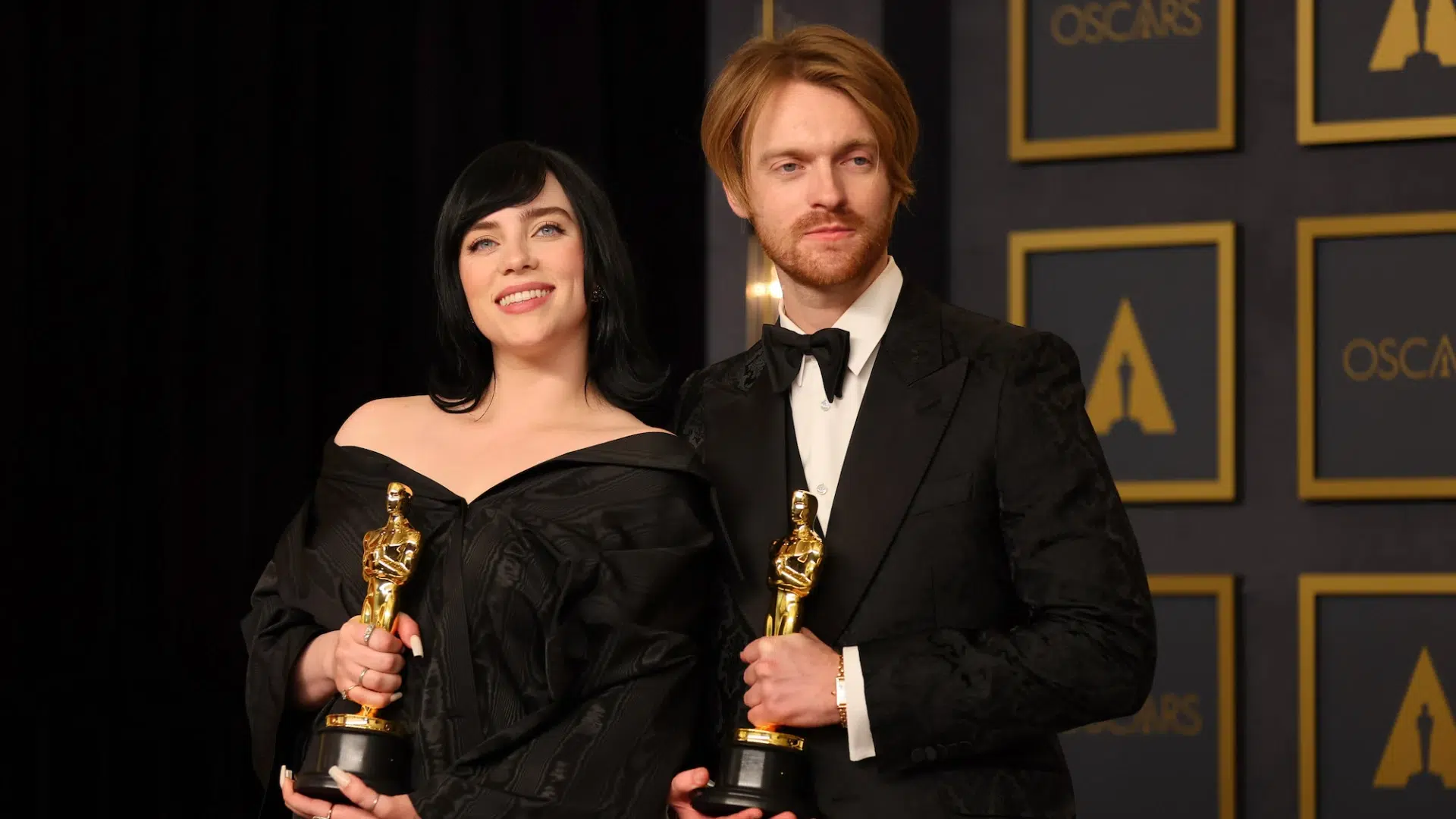 Billie Eilish and Finneas O'Connell smile while holding their Oscar awards against a backdrop of a gold Oscar statuette