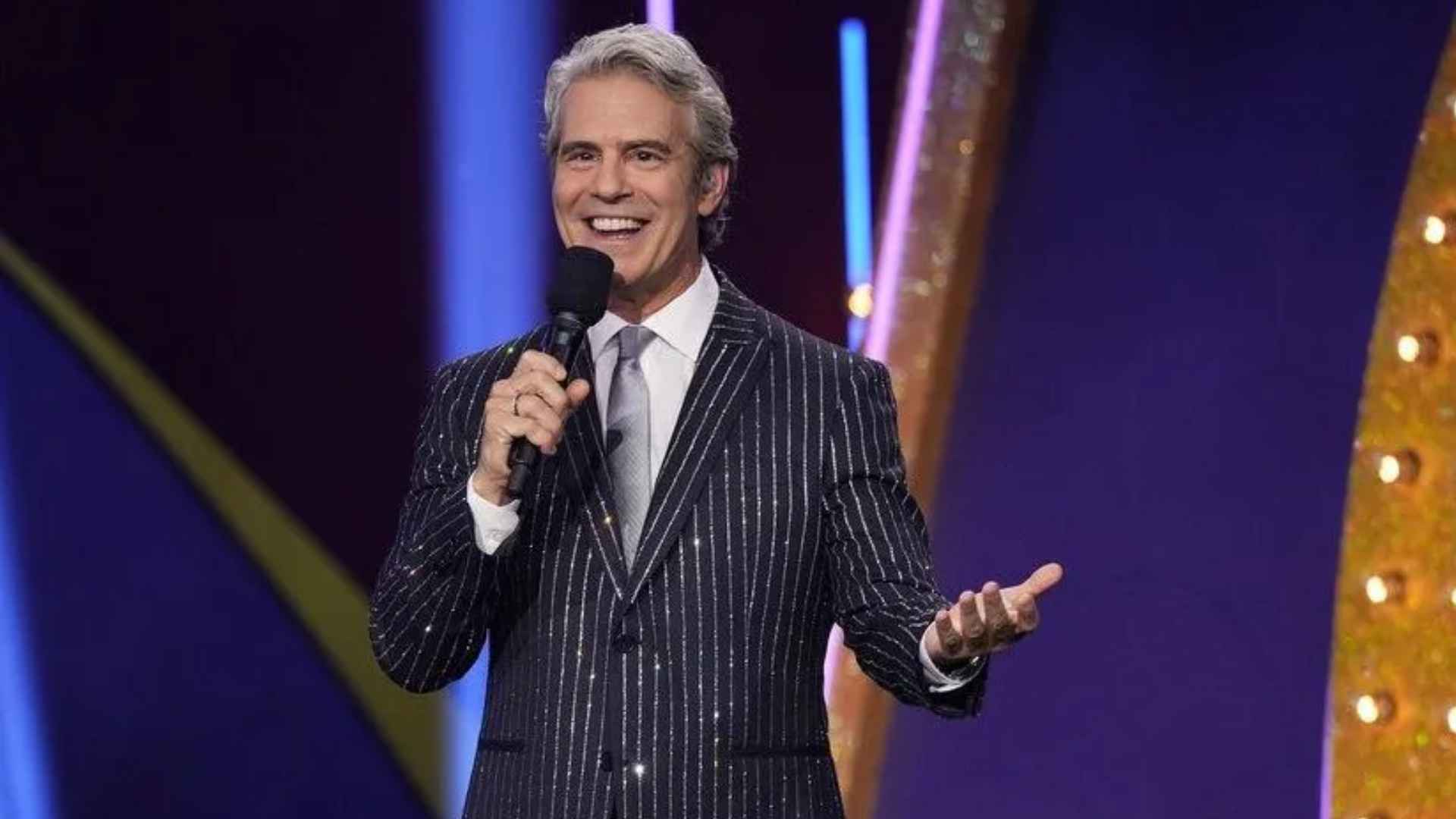 Andy Cohen smiling on stage in a pinstripe suit while holding a microphone during a live television event.