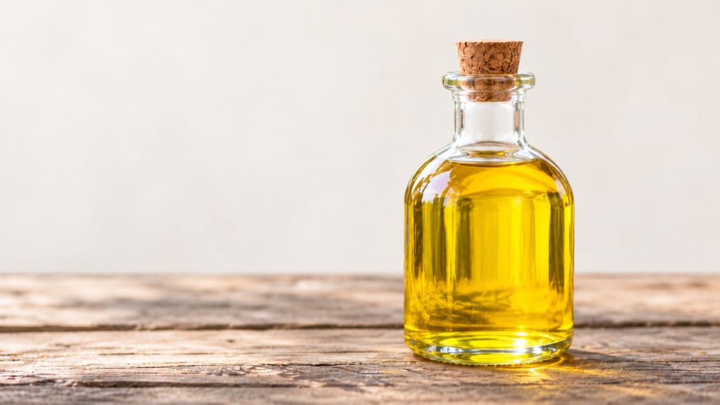 Clear glass bottle filled with golden canola oil on a rustic wooden surface against a plain beige background.