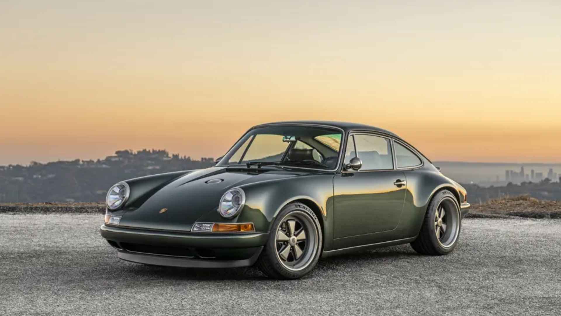 Dark green Porsche 911 reimagined by Singer, parked on gravel at sunset with a distant city skyline.