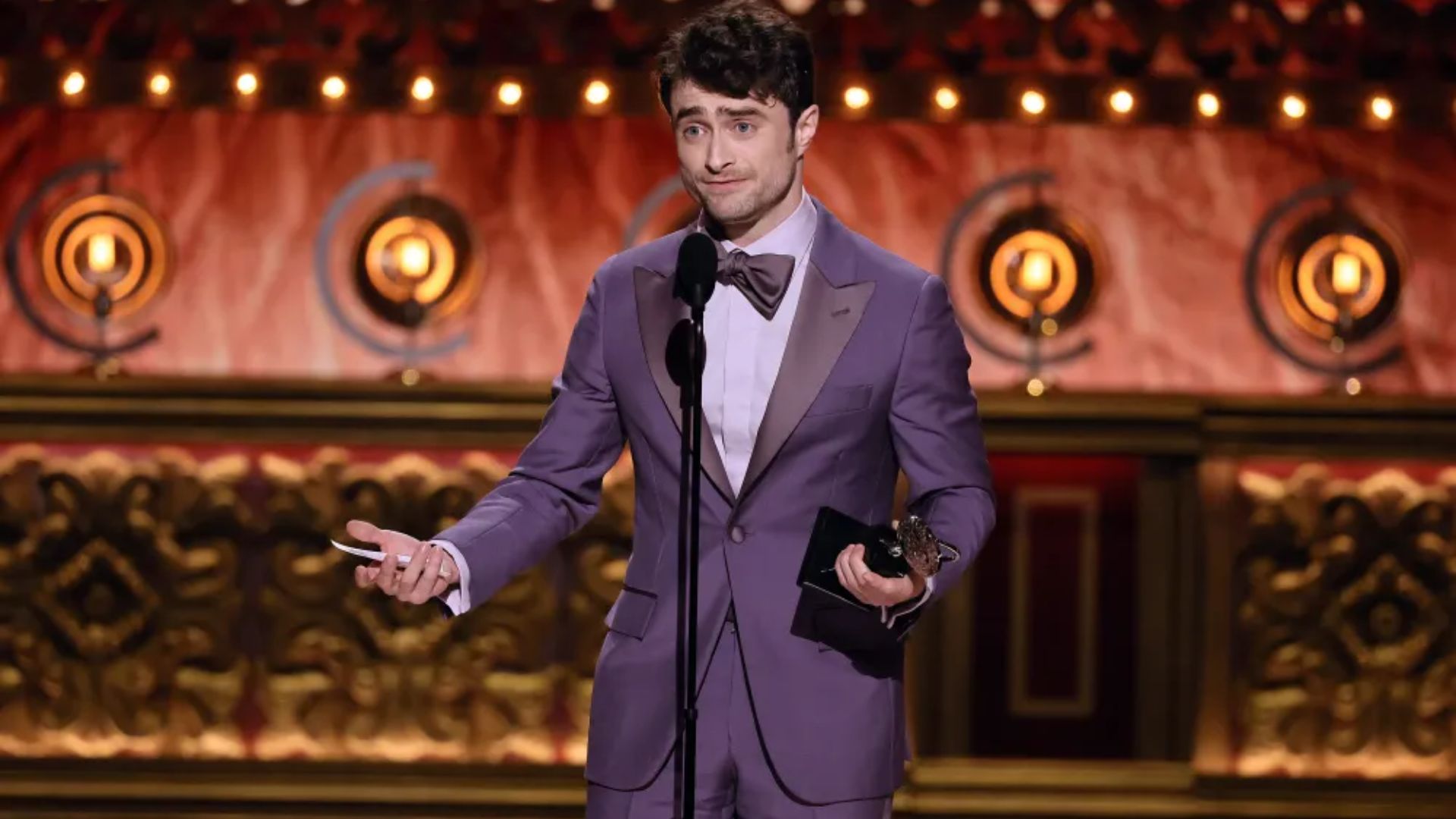Daniel Radcliffe accepting an award on stage while wearing a purple suit and bow tie