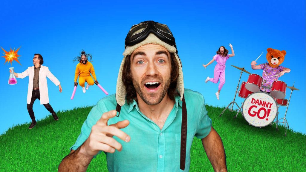 Danny Go performing in a colorful outdoor-style set with musical props and characters