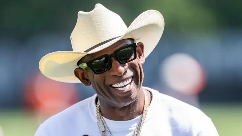 Deion Sanders smiling in a white cowboy hat and black sunglasses, wearing a white shirt and gold chains outdoors.