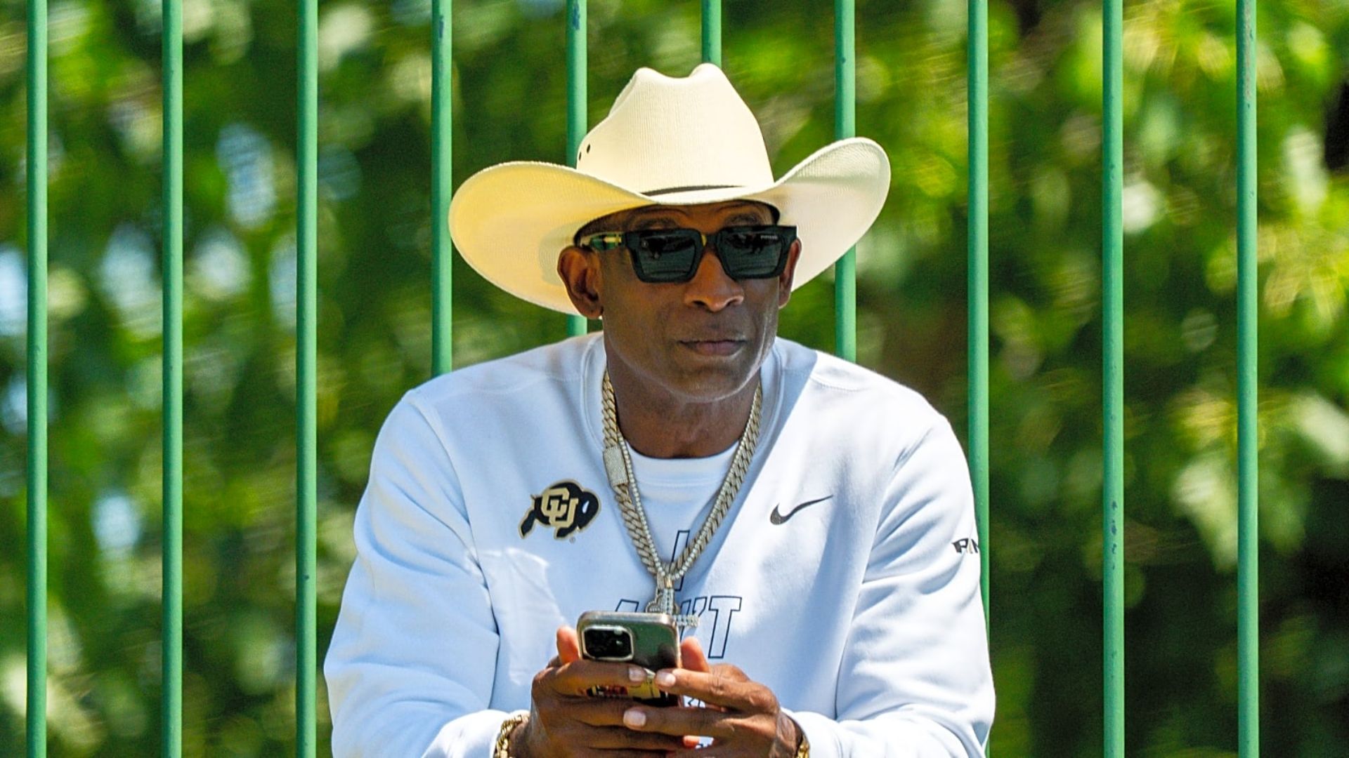 Deion Sanders wearing a white cowboy hat and black sunglasses, holding a phone while seated outdoors by a green fence.