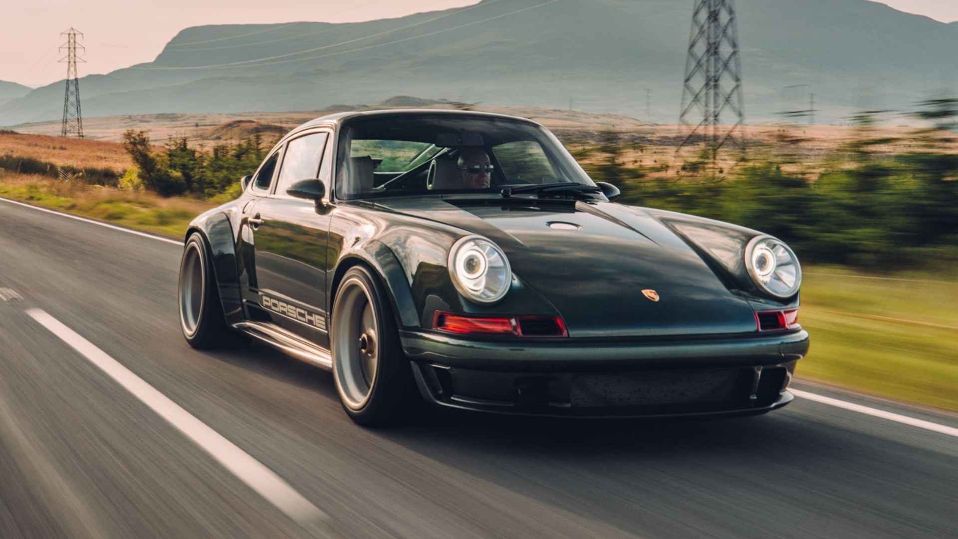 Dark green Singer DLS Porsche 911 driving on a rural road with mountains and fields in the distance.