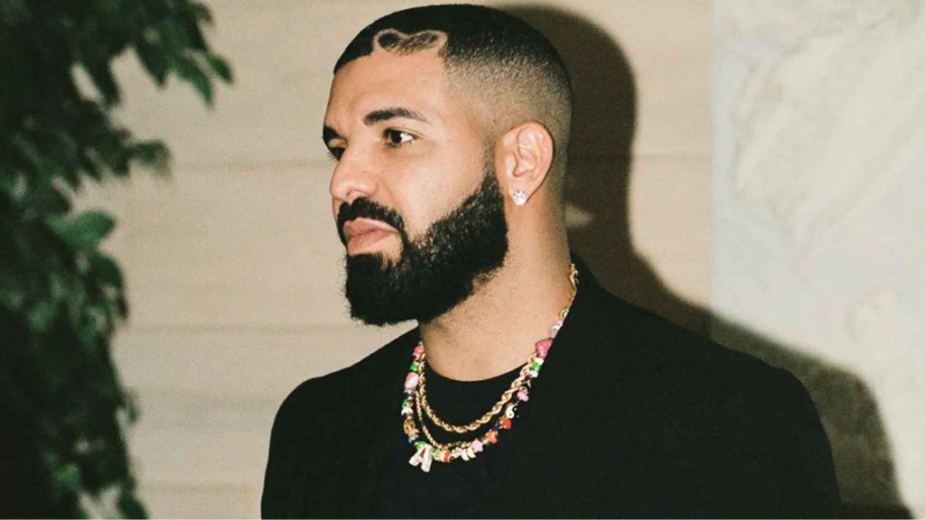 Drake wearing a black shirt and layered necklaces while posing indoors with a neutral background.