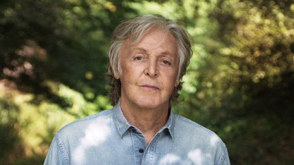 Paul McCartney standing outdoors in a light blue shirt with a softly blurred green garden background.