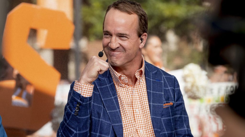 Peyton Manning wearing a blue checked blazer and orange shirt, smiling during an outdoor sports broadcast.