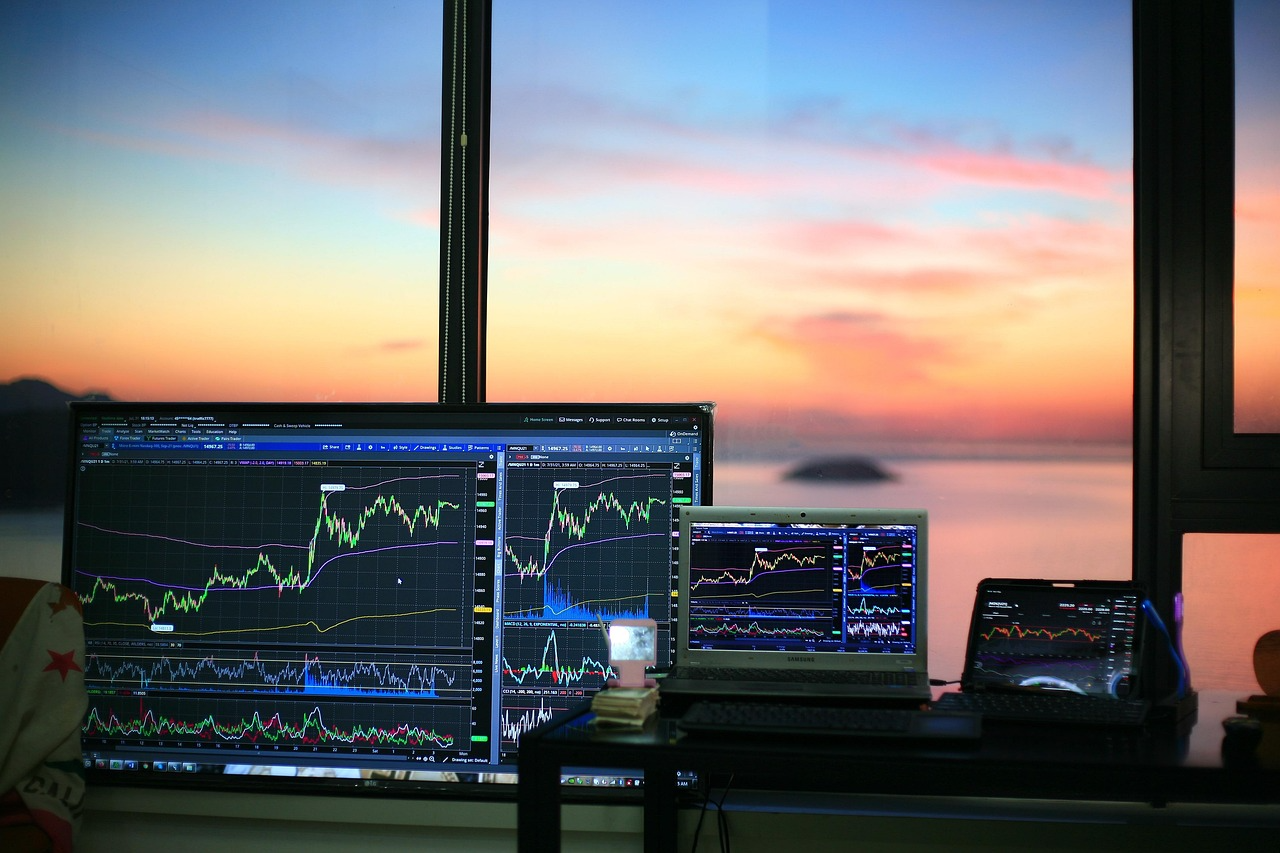 Multiple screens displaying stock market graphs in an office setting with sunset view