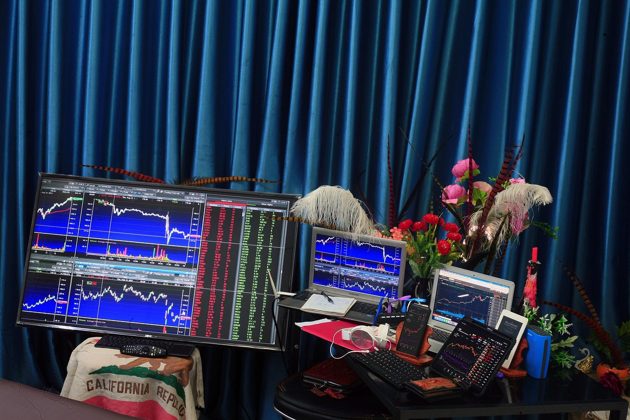 Multiple monitors displaying stock market charts with vibrant floral arrangement against blue curtains