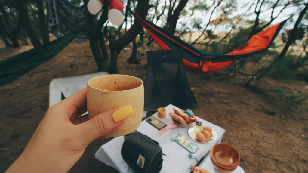 Hand holding wooden cup in forest campsite with hammock and picnic setup on table