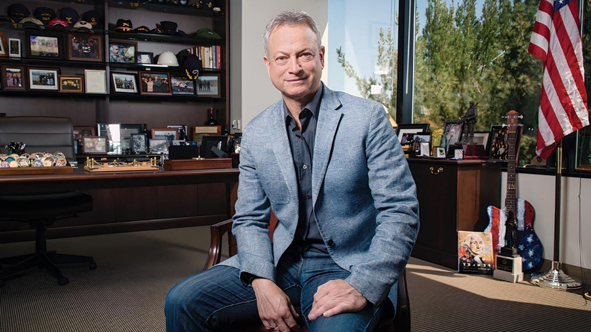 Gary Alan Sinise sitting in an office with framed photos, guitars, and an American flag in the background.