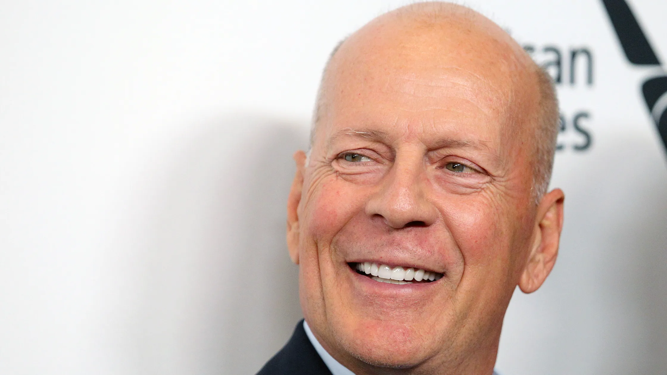 close-up of bruce willis smiling in formal attire against a white backdrop during a public event