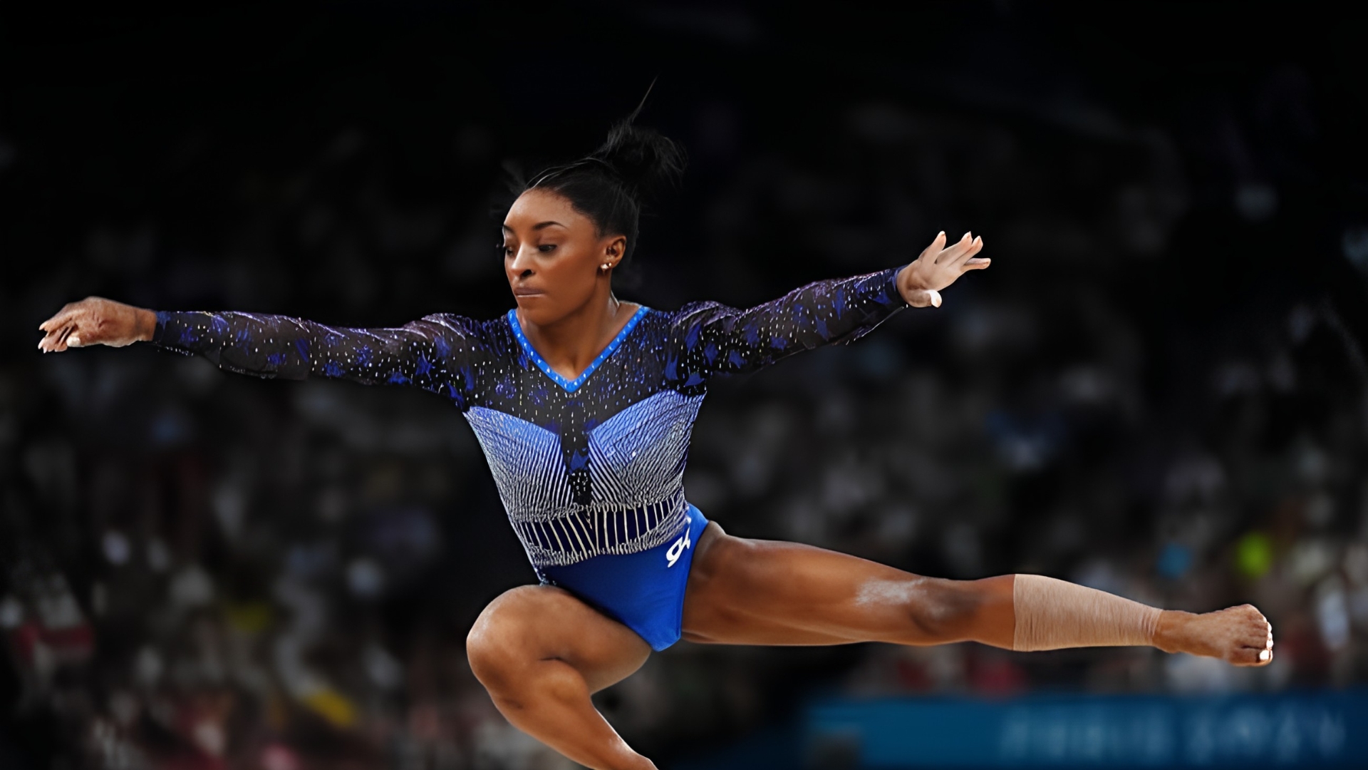Simone Biles balancing on a balance beam in a blue and black embellished leotard at the Paris 2024 Olympics.