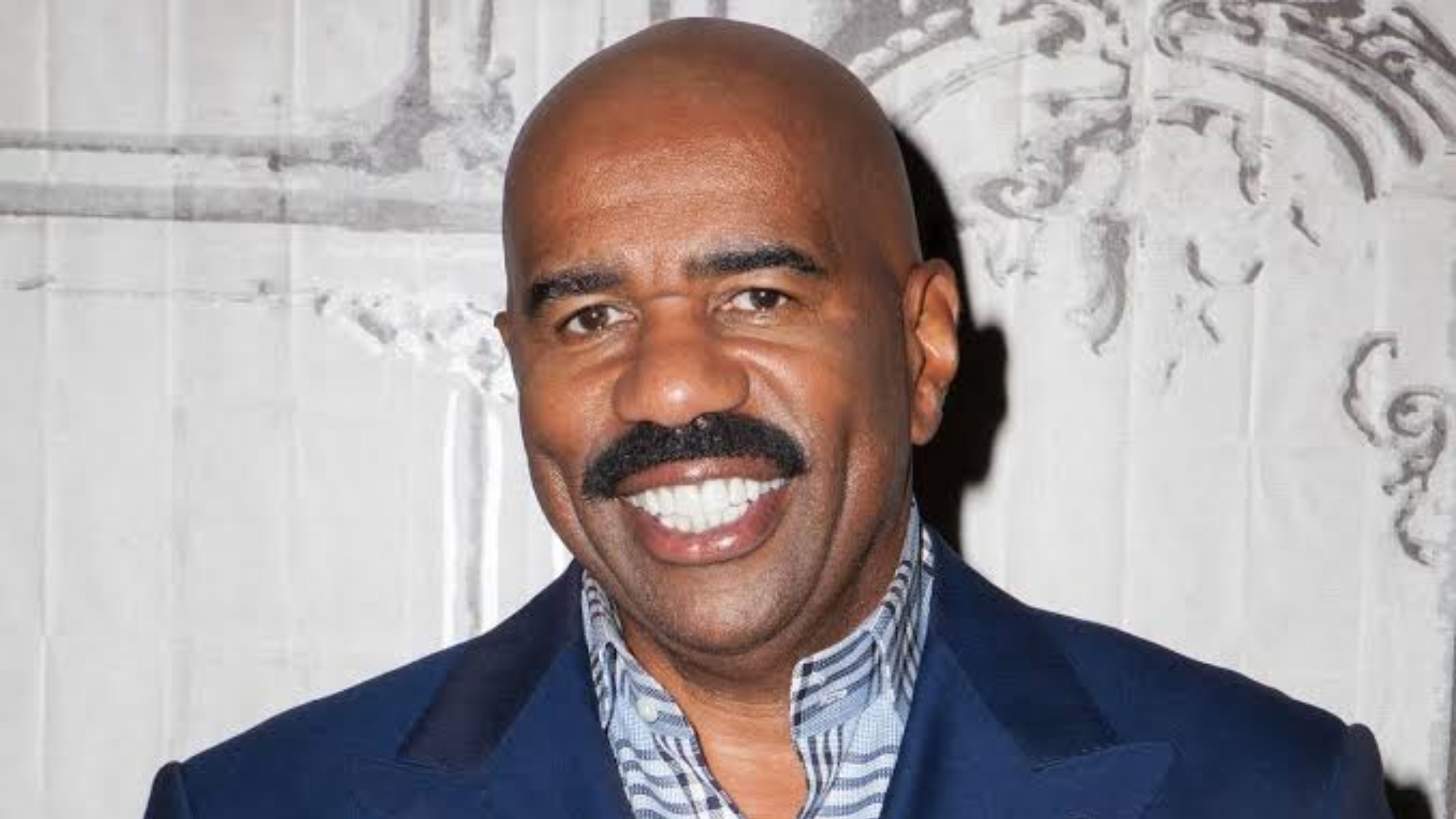Close-up headshot of Steve Harvey smiling in a blue suit jacket and patterned shirt.