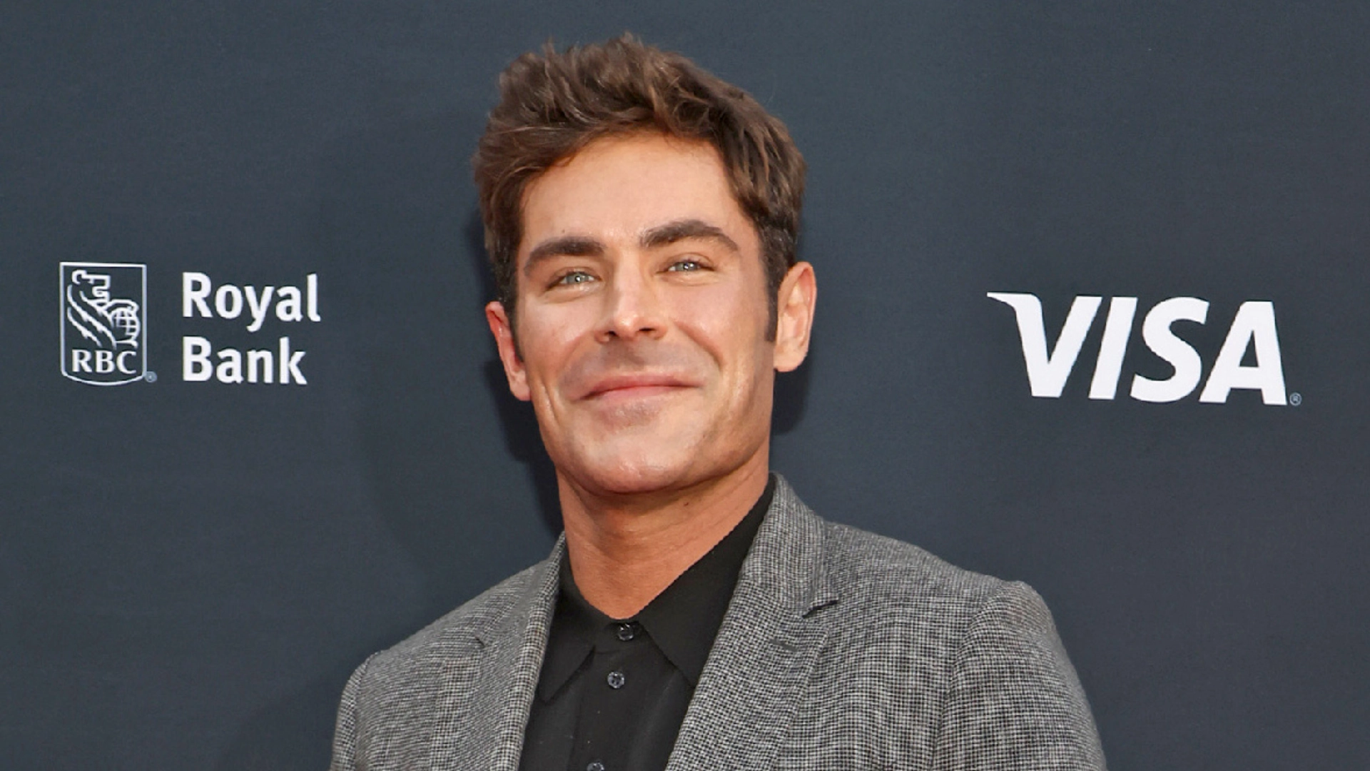 Zac Efron wearing a gray checkered suit and black shirt while posing against a dark event backdrop